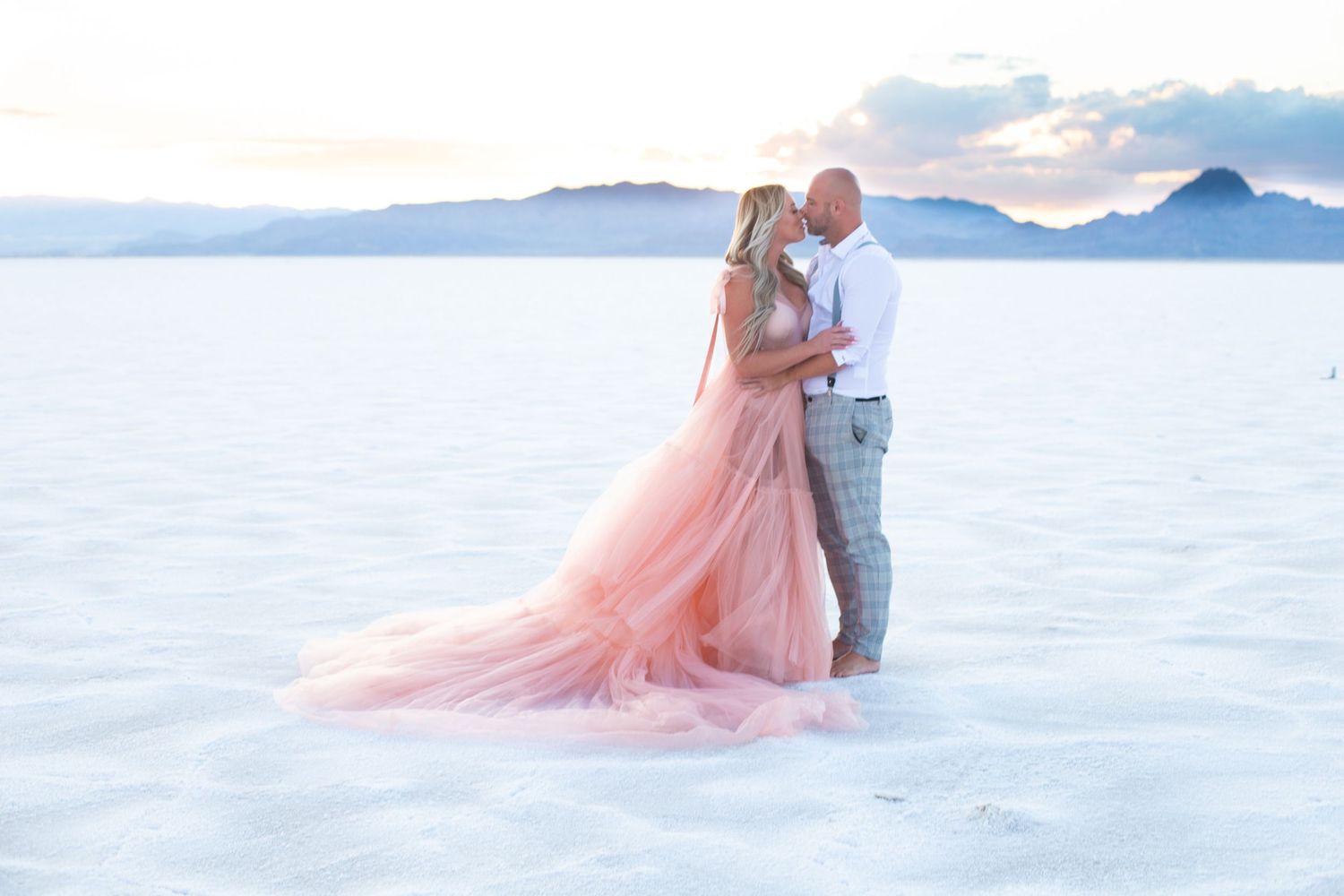 Couple shares intimate moment on white sand beach with pink gown flowing in breeze.