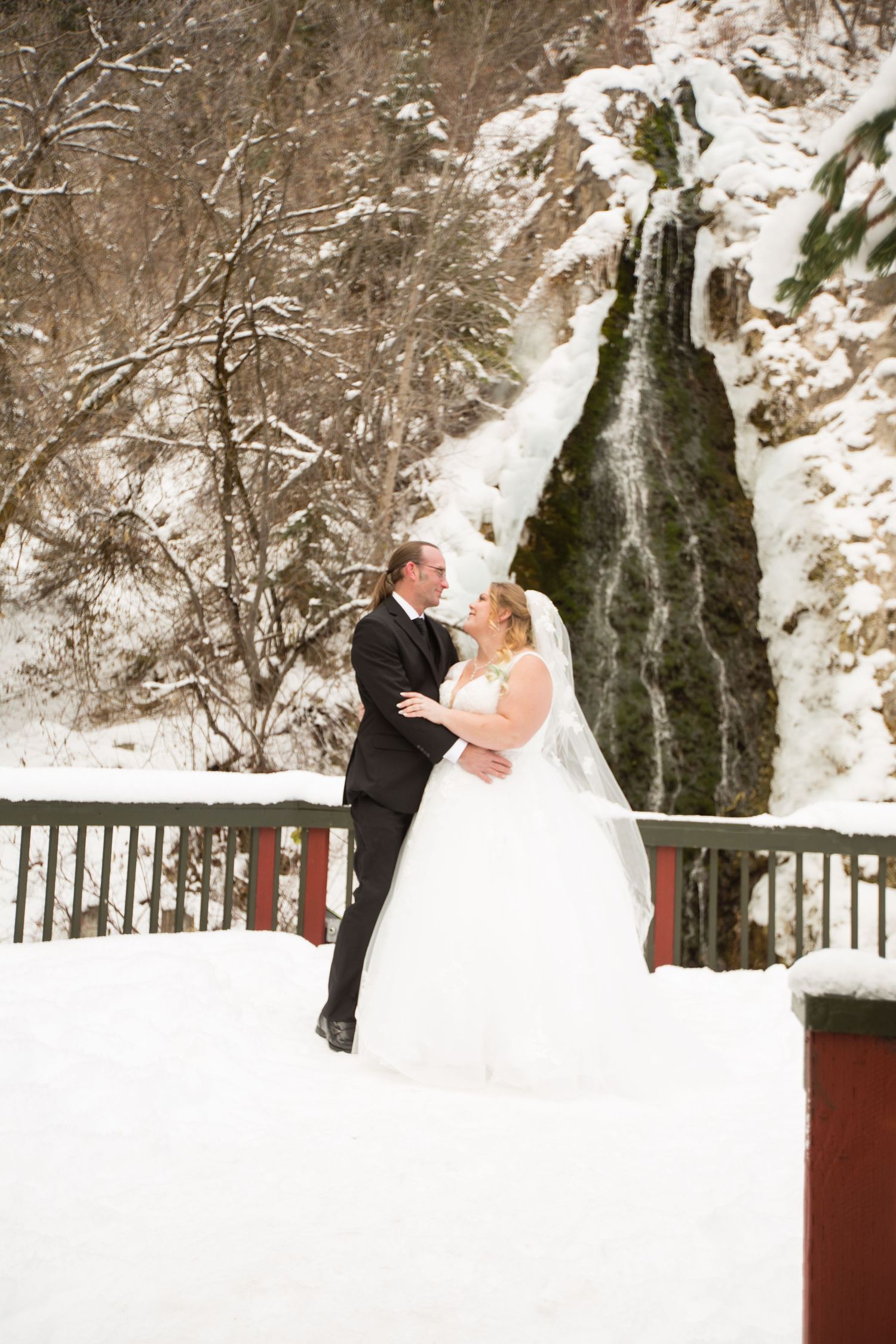 Newlyweds embrace on snowy bridge platform with frozen waterfall cascading behind them.