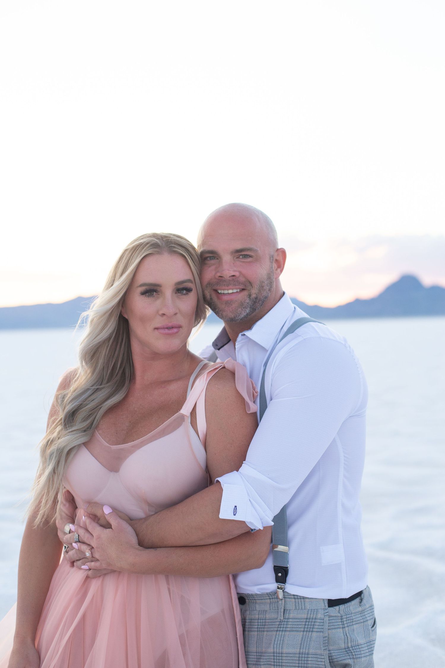 Romantic couple embraces on a beach at sunset with mountains in background.