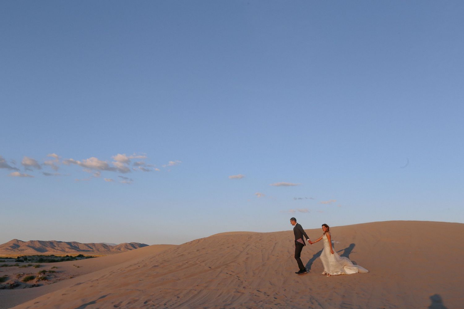 A romantic couple poses for photos across dramatic sand dunes in the desert at sunset.