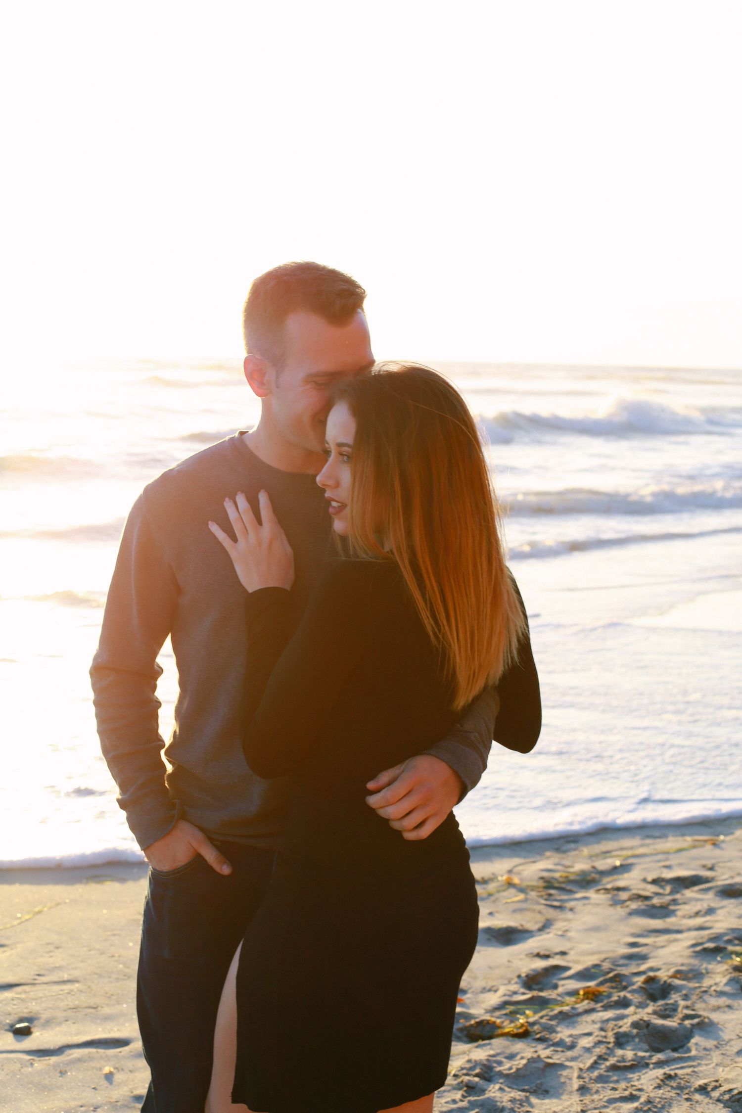Couple embracing on beach at sunset with ocean waves in background.