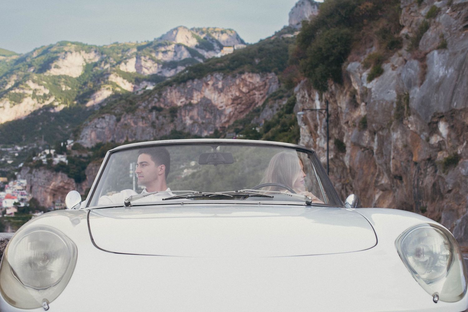 Couple riding in a classic white convertible sports car along a scenic Amalfi Coast mountain road.