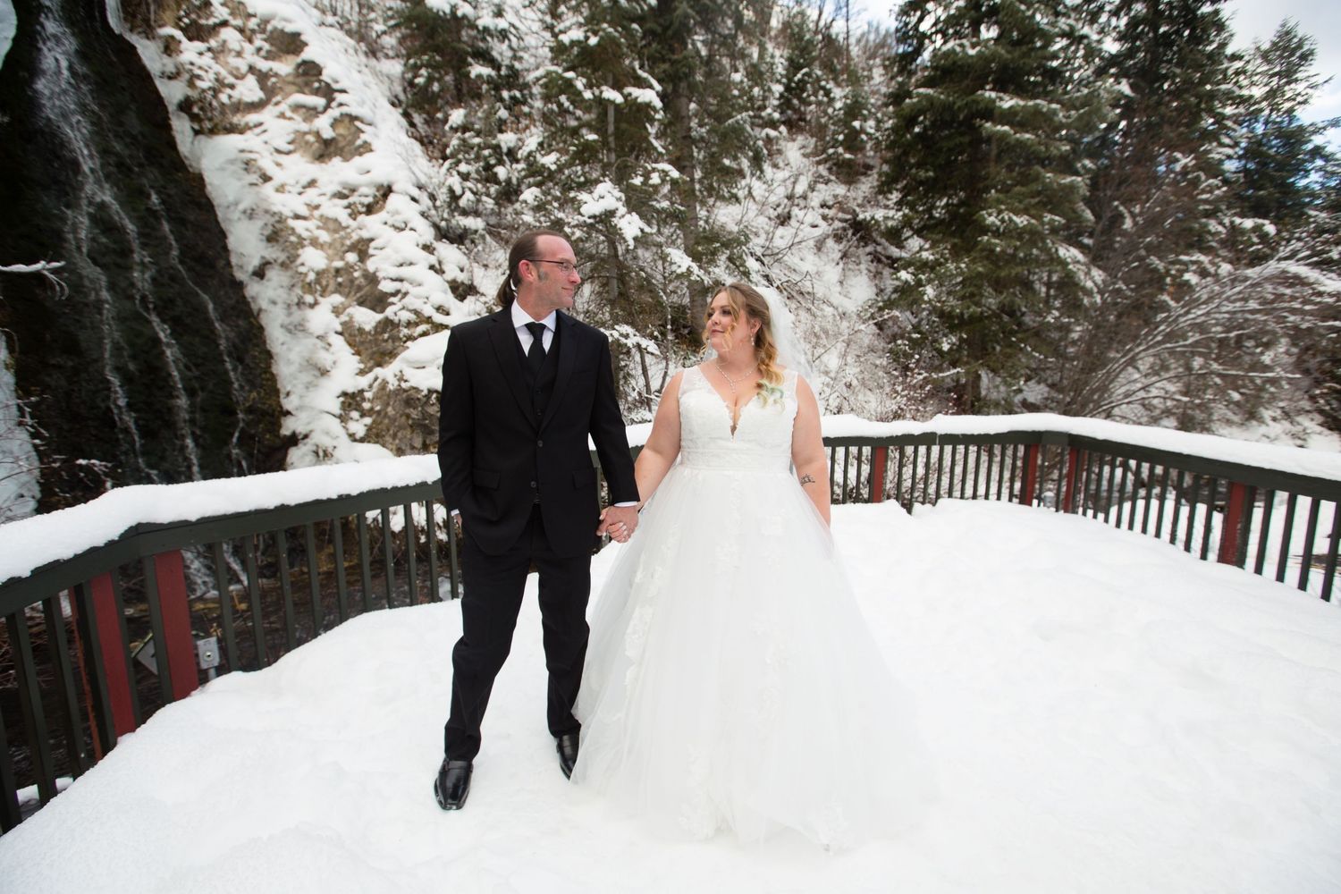 Wedding couple strolls along a snow-covered wooden walkway in a winter forest setting.