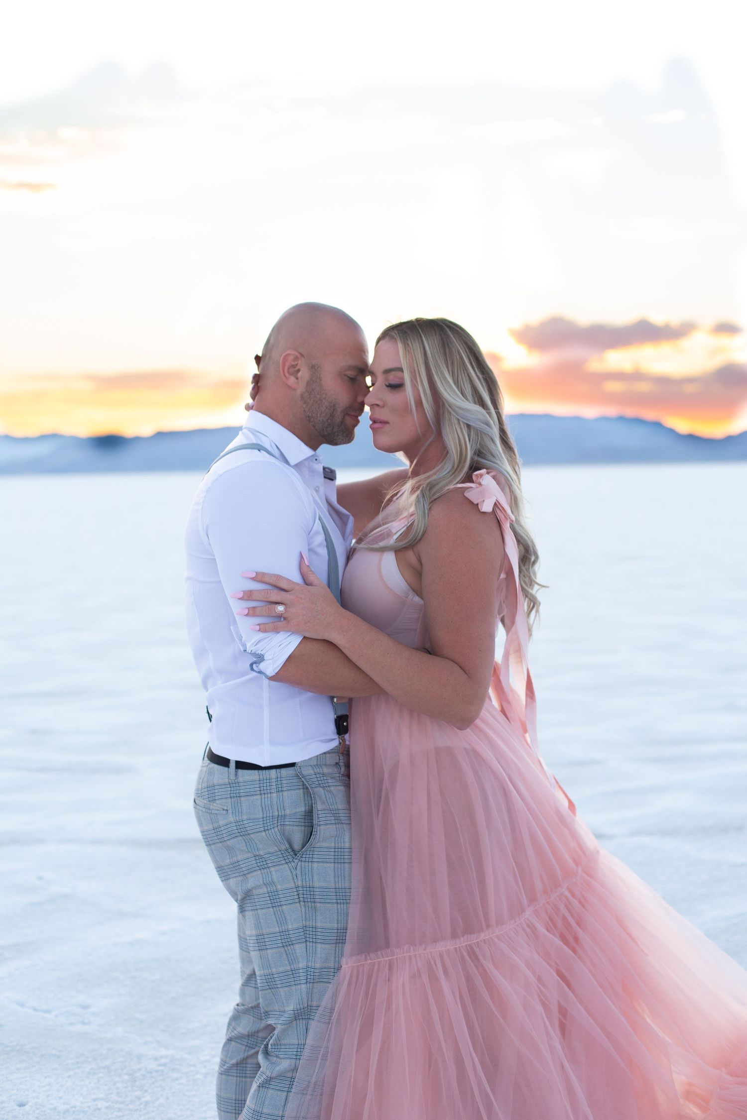 Couple shares an intimate moment on white salt flats with mountains and sunset in background.