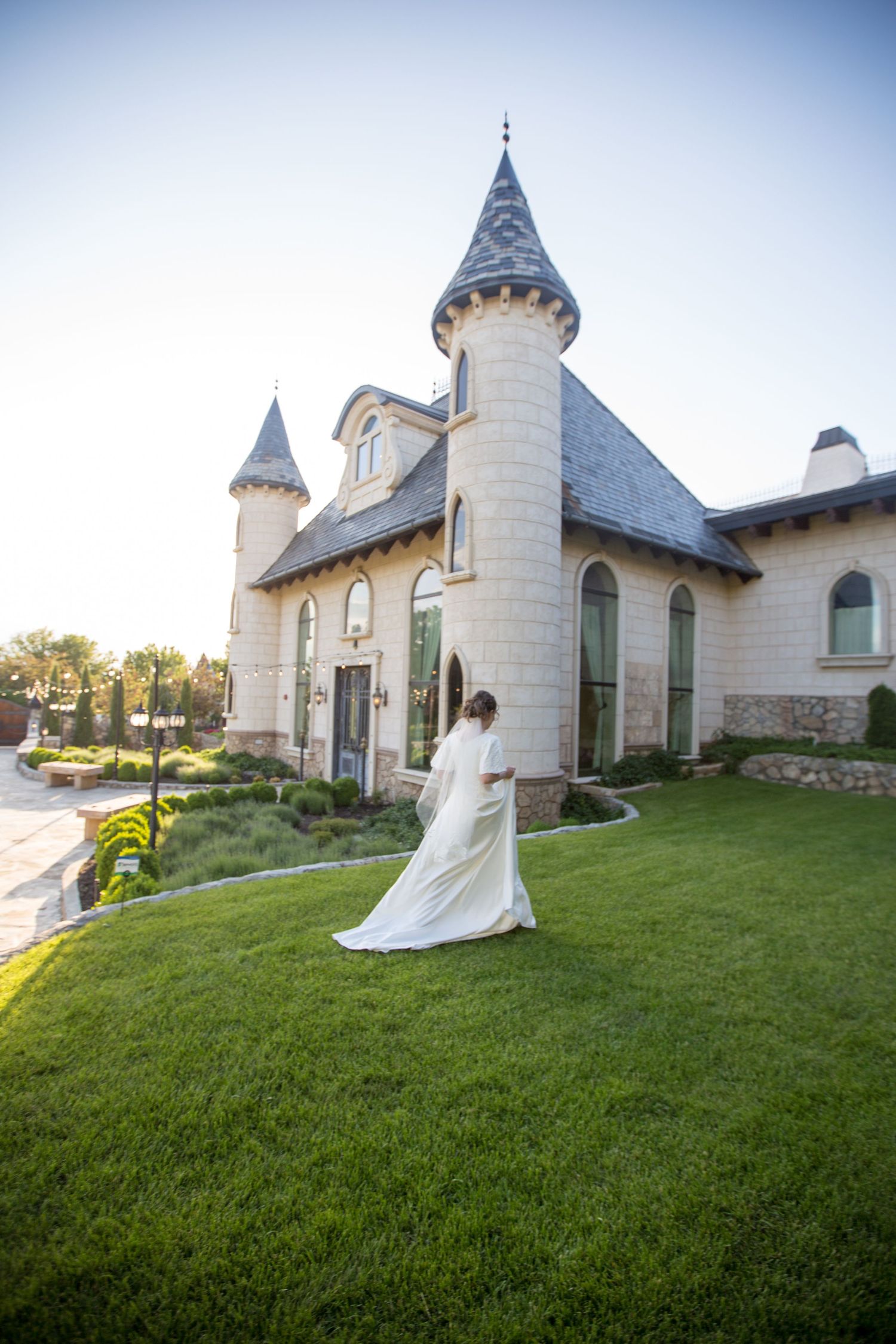 Bride in flowing white dress twirls on green lawn in front of castle-like wedding venue.