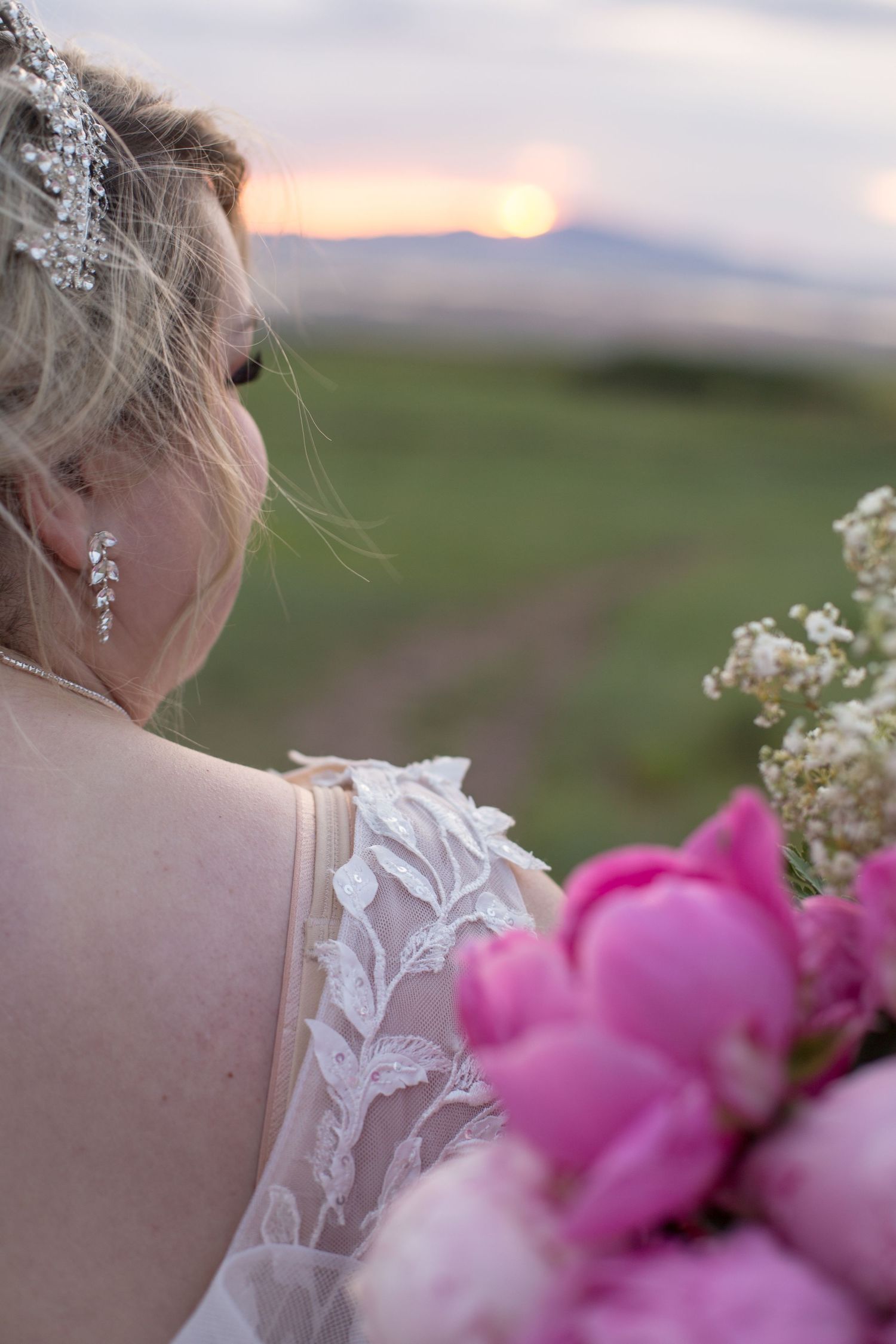 Lace wedding dress detail and pink flowers against a sunset mountain backdrop.