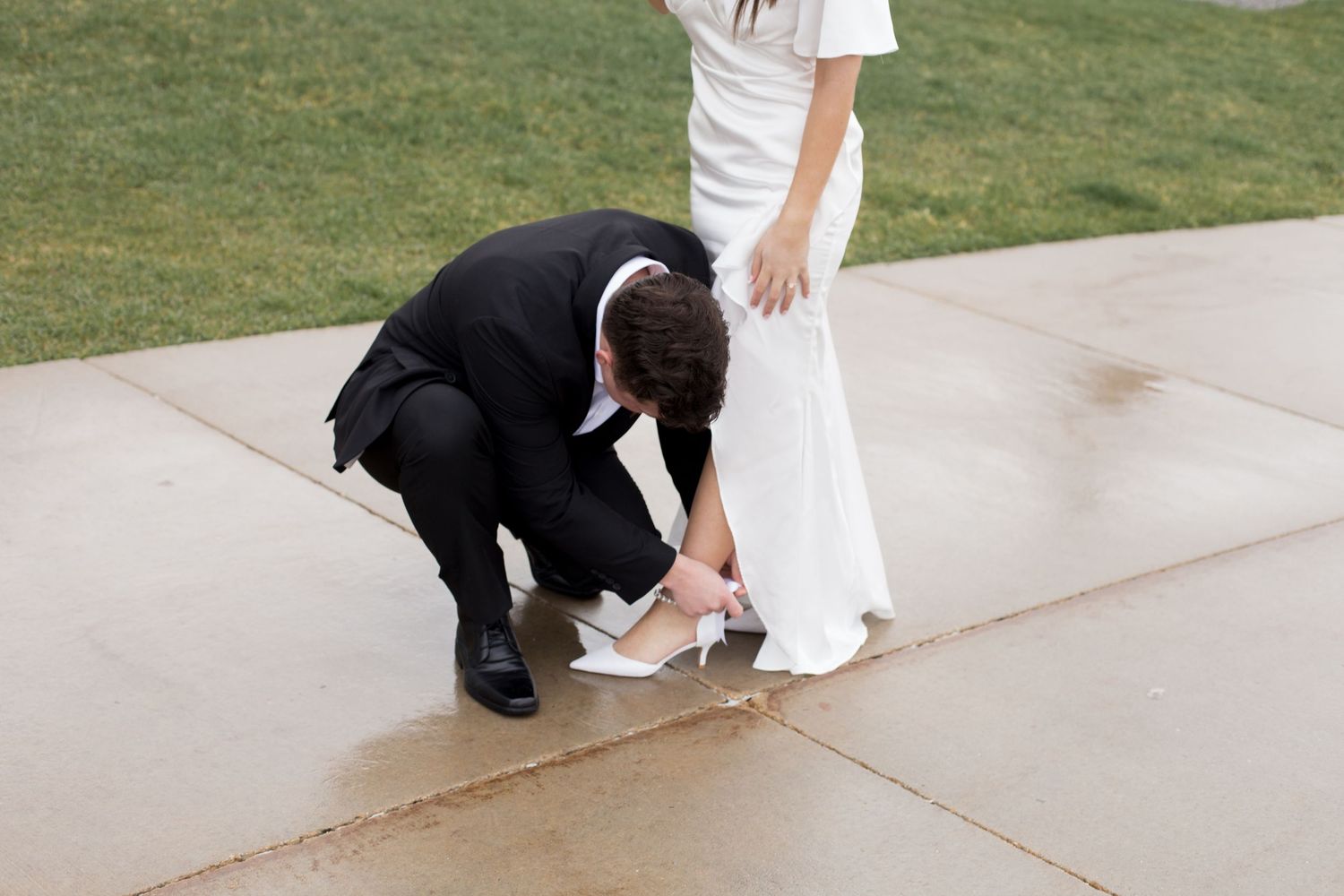 A couple in formal wear poses on a grassy lawn near a white building.