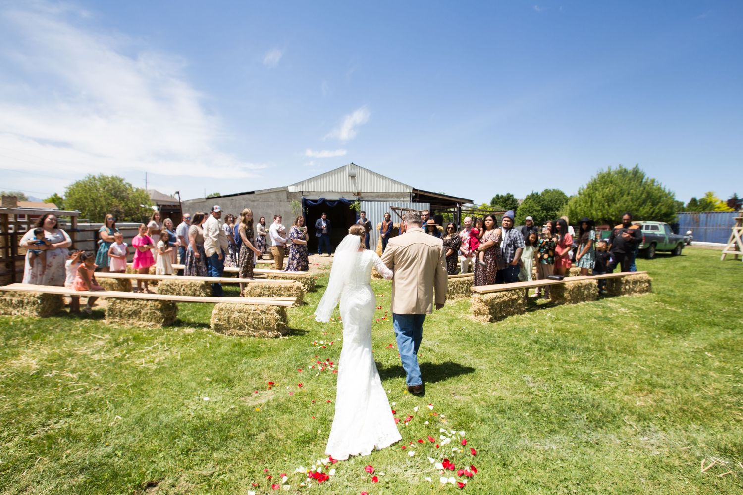 Bride and groom walk down outdoor aisle lined with hay bales at rustic farm wedding on sunny day.
