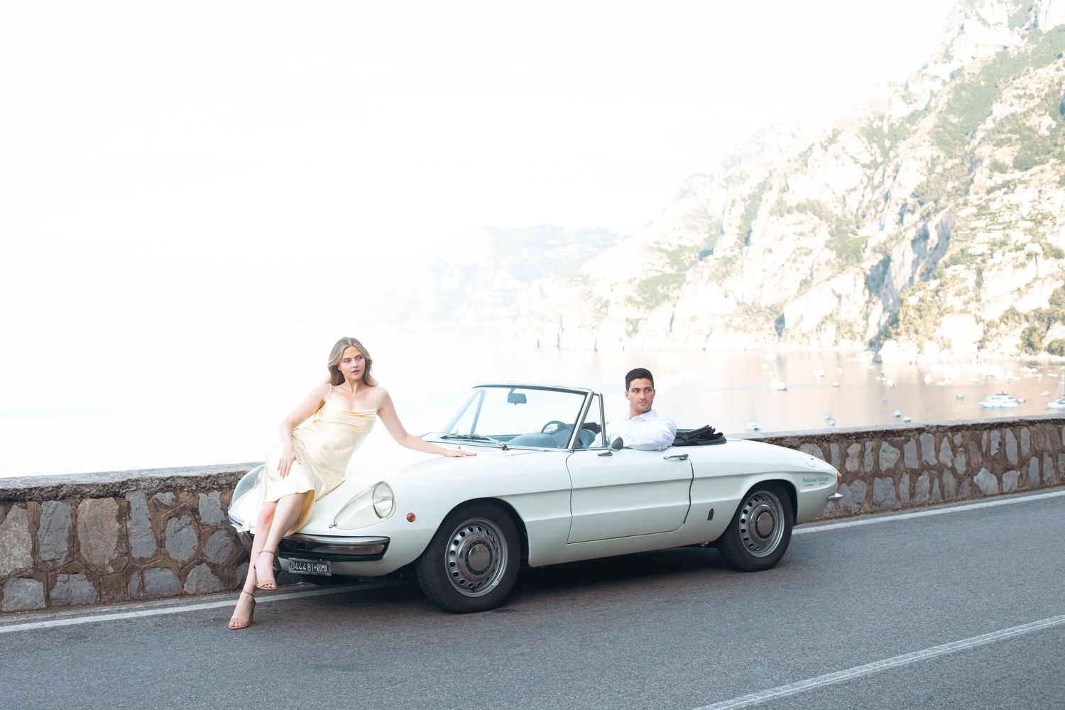 Woman in yellow silk dress posing with man beside white classic Alfa Romeo Spider on Amalfi Coast.