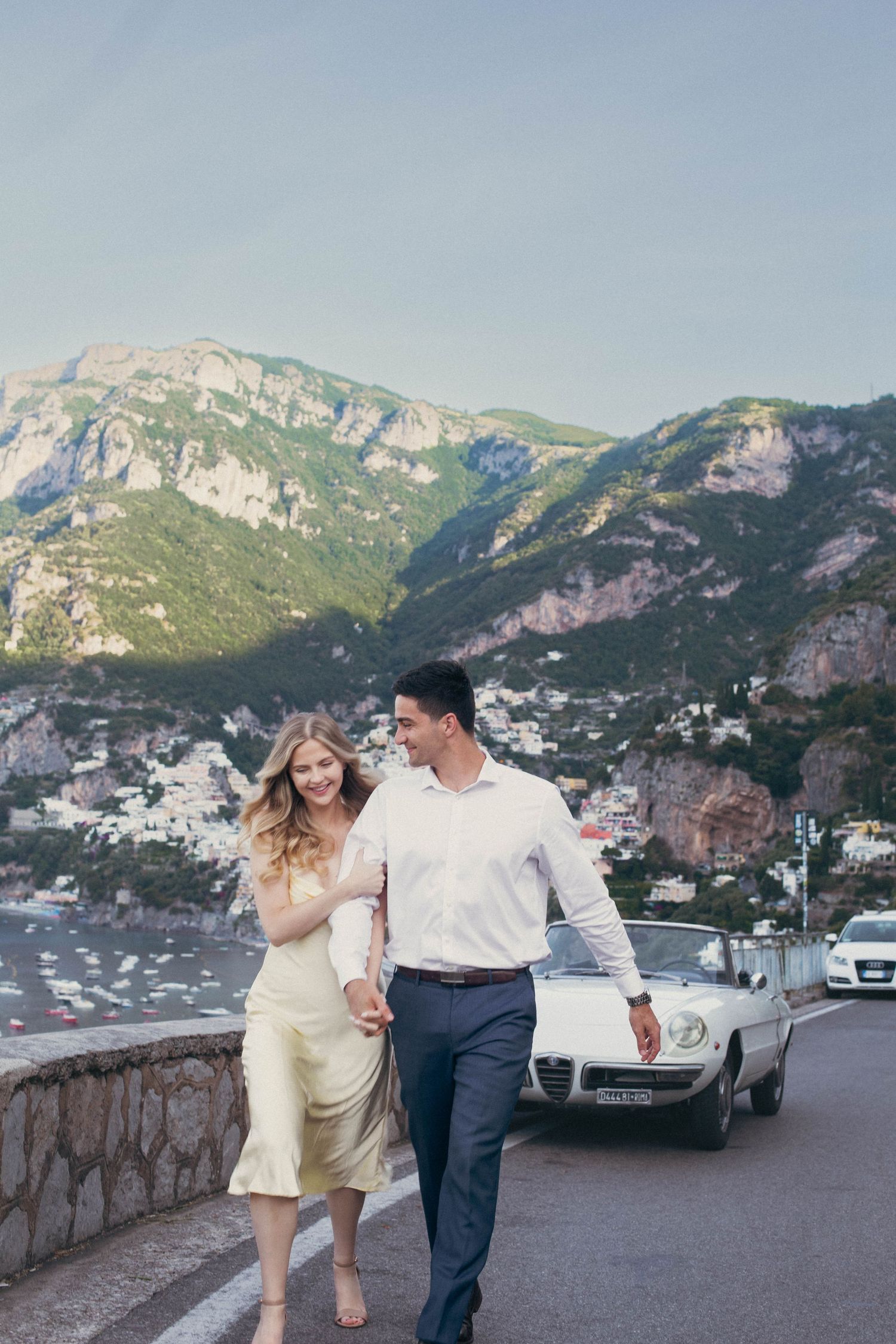Couple walking hand in hand on Amalfi Coast road with white vintage Alfa Romeo and Positano hillside behind.