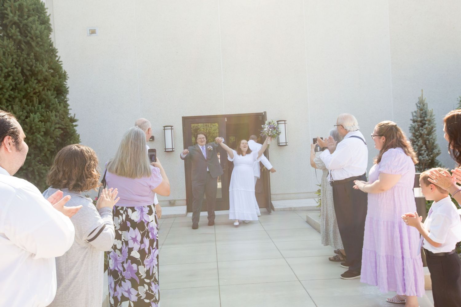 Newlywed couple exits wedding venue as guests celebrate and applaud outside a modern building.