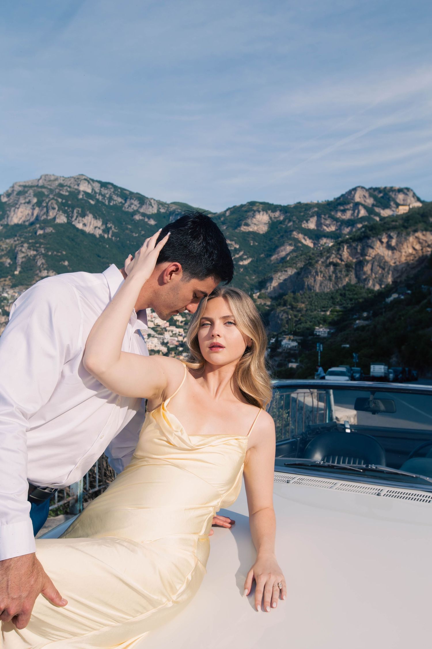 Elegant couple in yellow dress and white shirt posing on yacht with Amalfi Coast mountains behind.