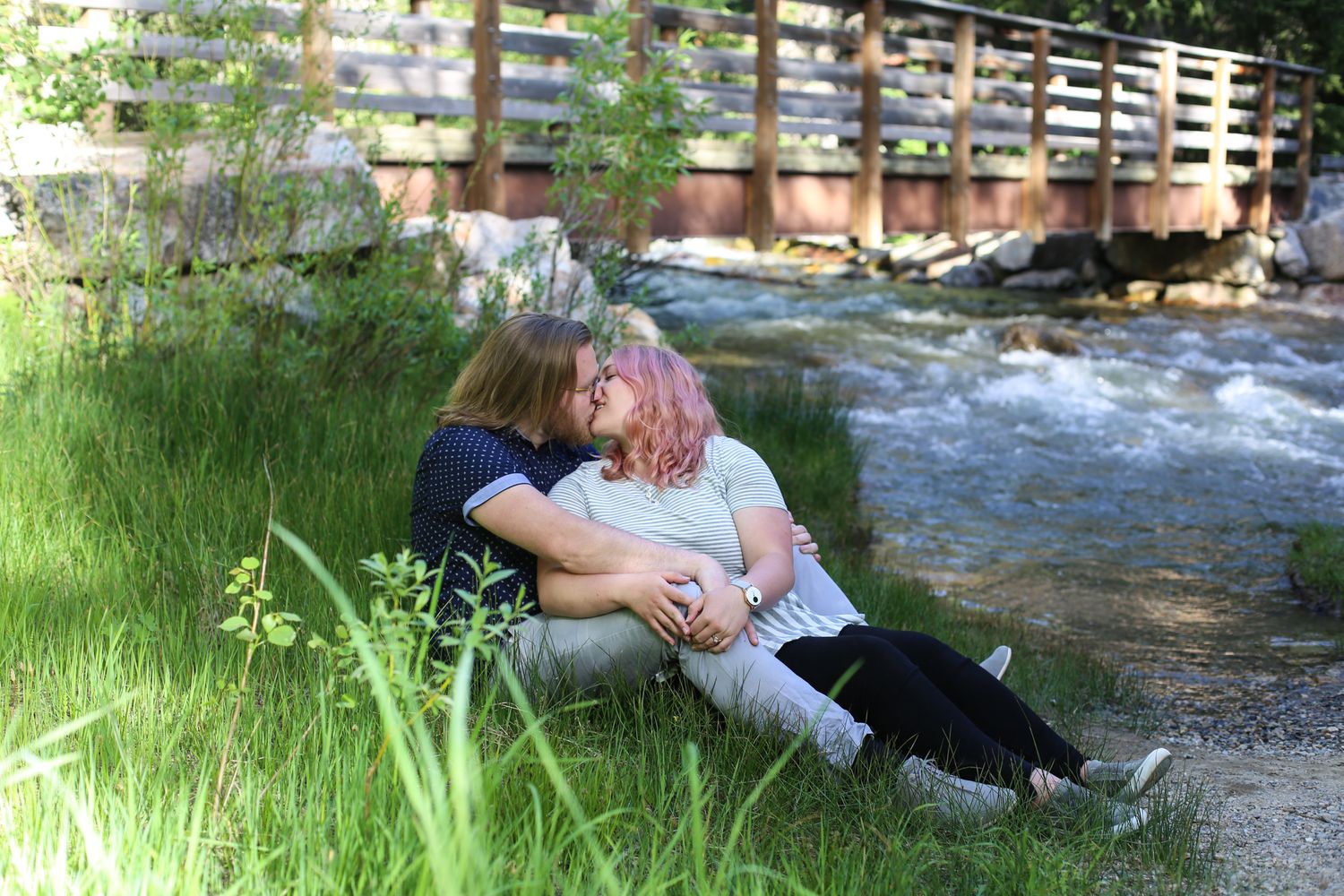 Couple sitting by a stream in lush green grass, sharing a romantic moment near a wooden bridge.