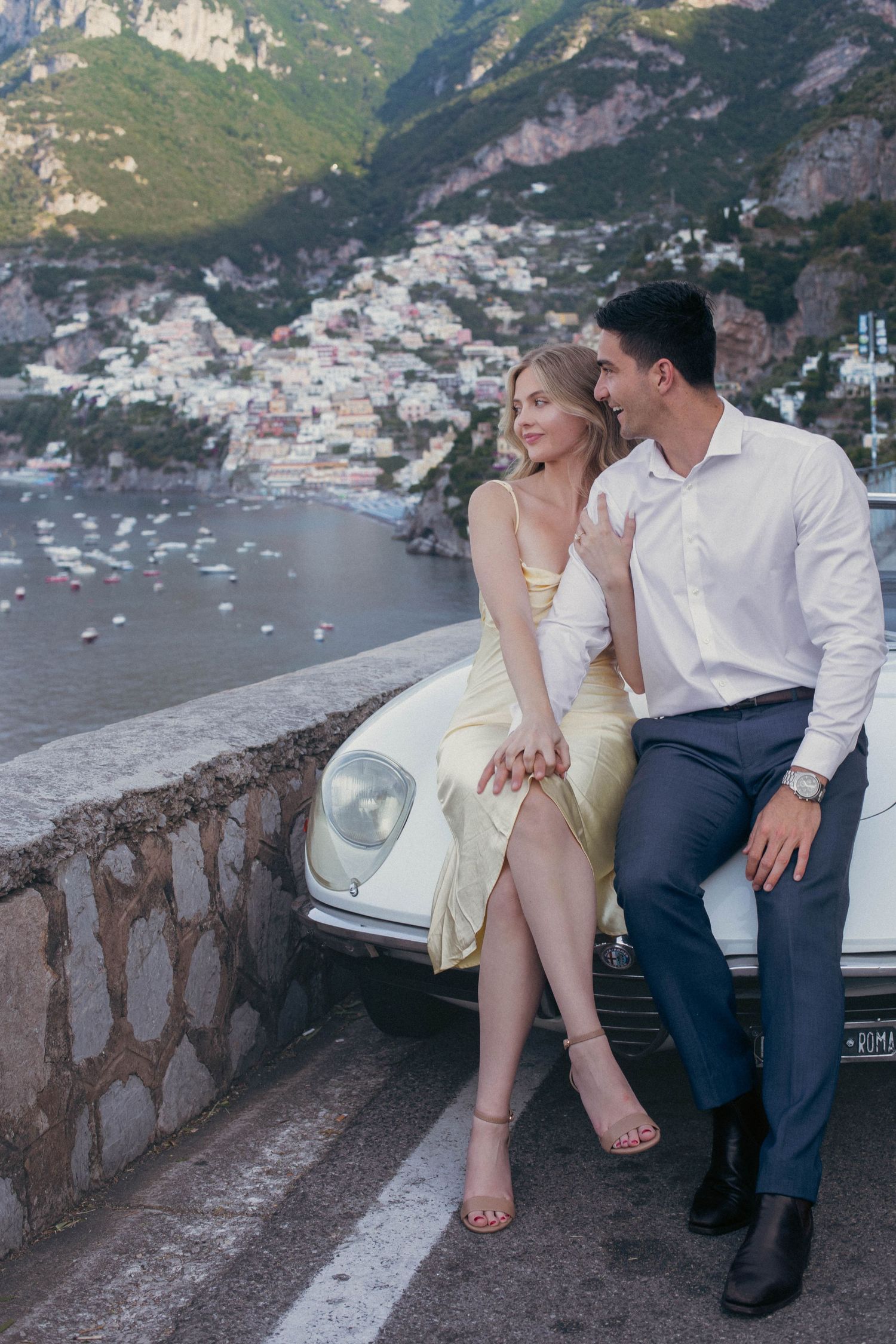 Romantic couple sitting on vintage white car hood with stunning Positano hillside village and sea in background.