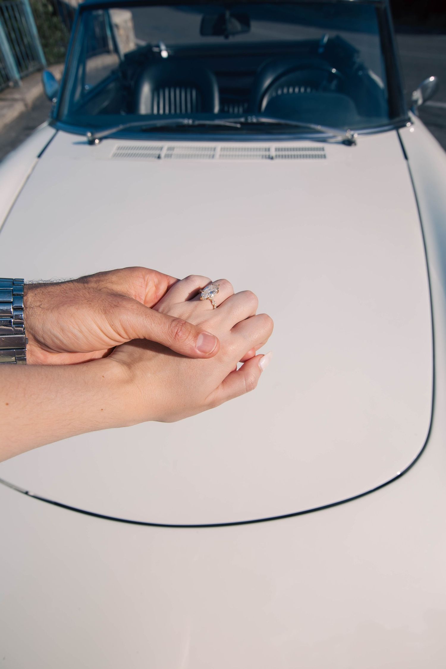 Close-up of couple holding hands with engagement ring on the hood of a vintage white convertible.