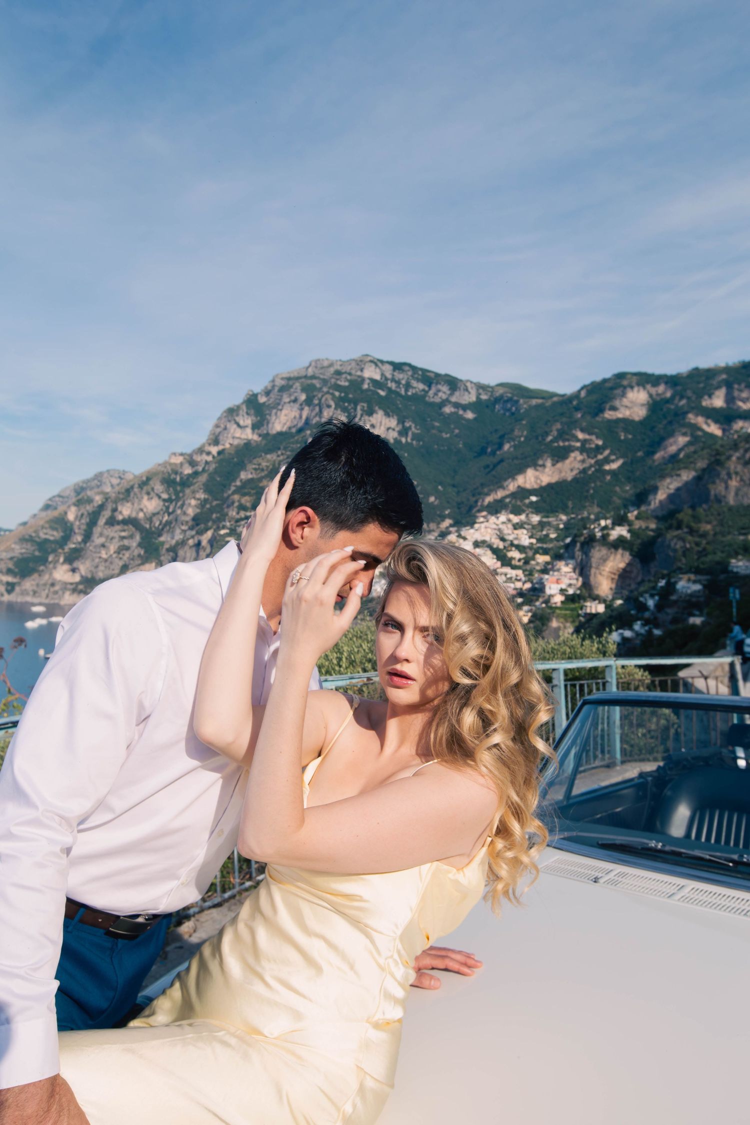 Couple sharing intimate moment leaning against white convertible with Positano mountains and blue sky.