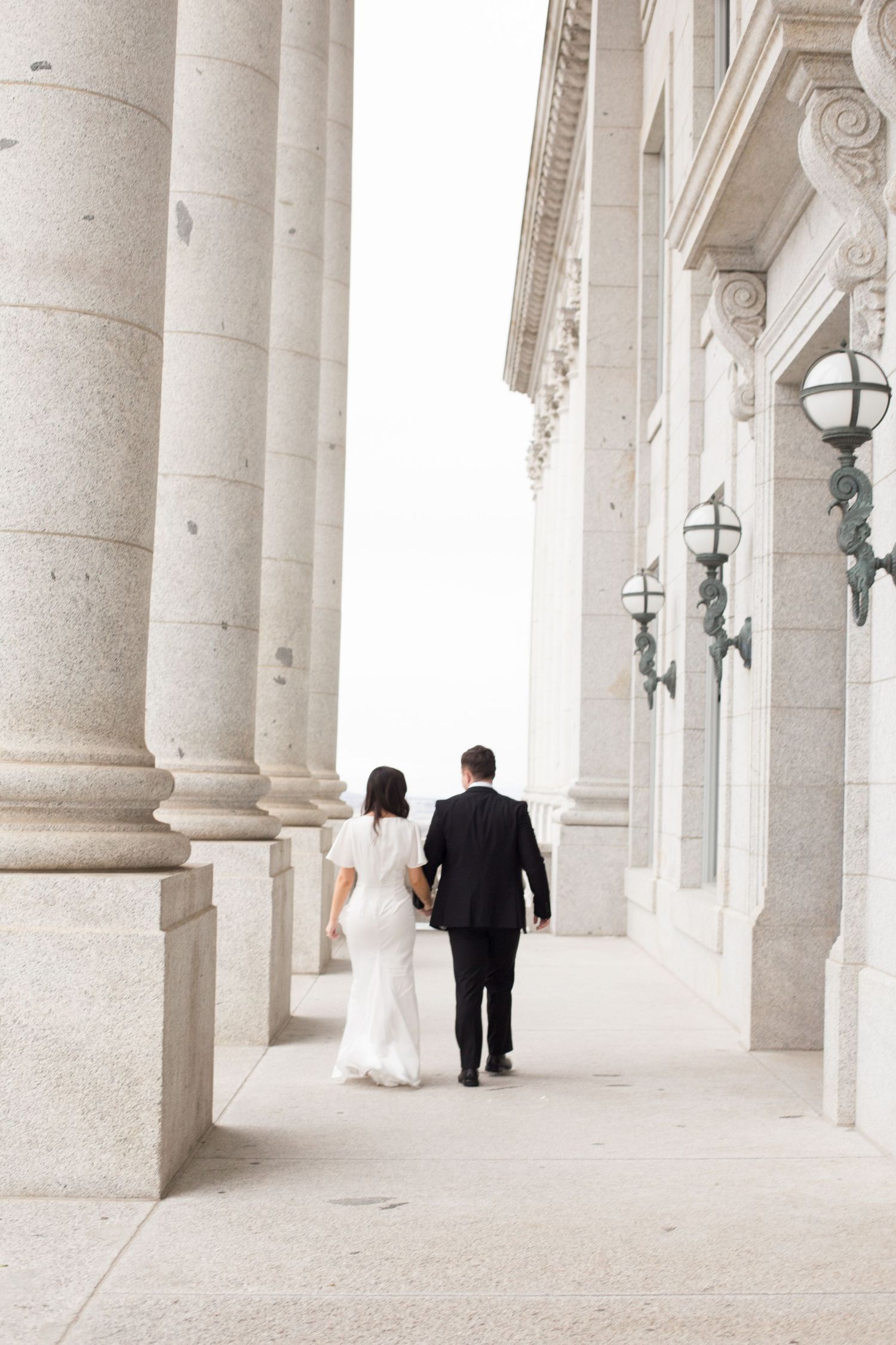 A couple in wedding attire walking through an elegant colonnade with stone pillars and vintage light fixtures.