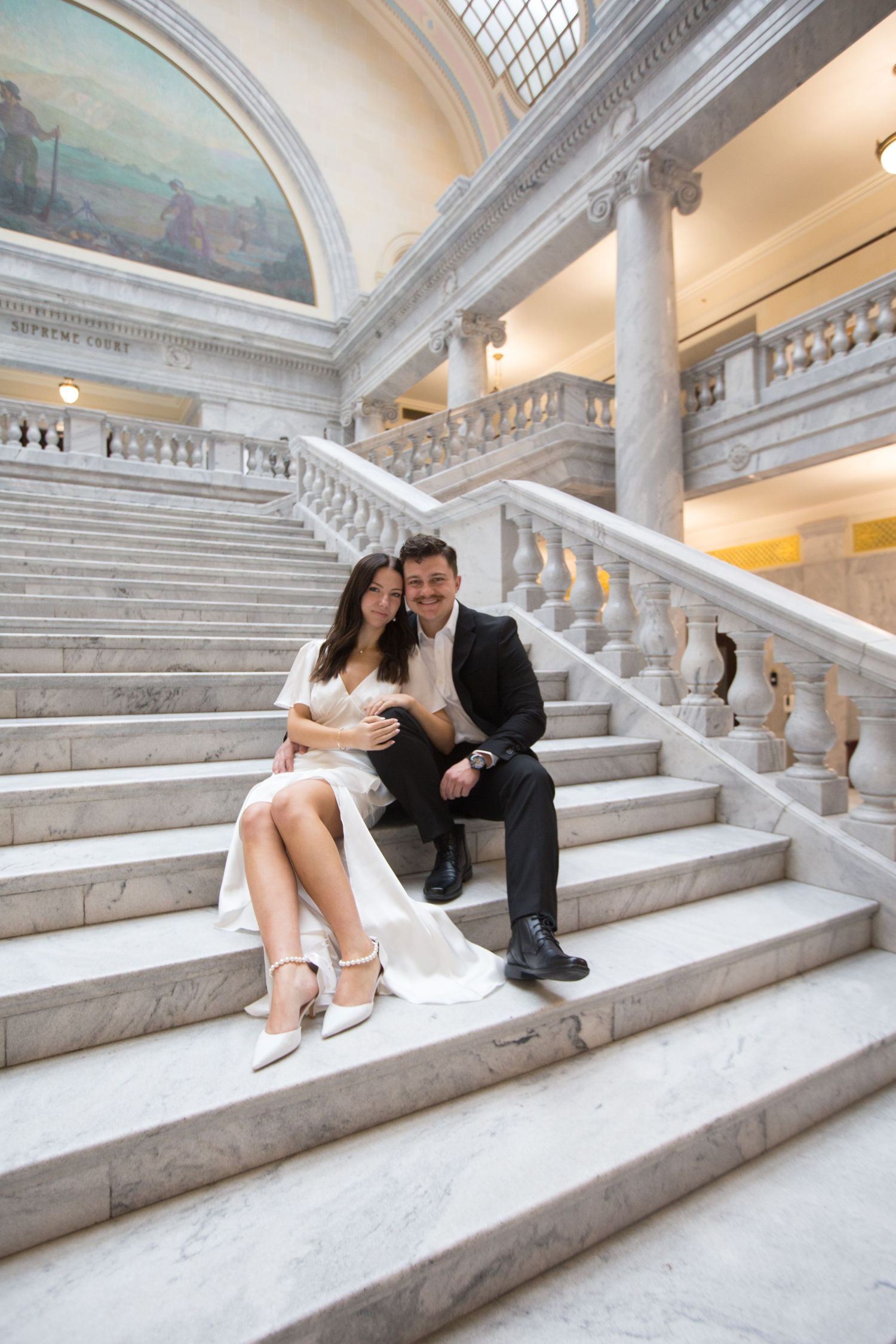 A couple sits together on elegant marble stairs beneath a painted dome and classical columns.