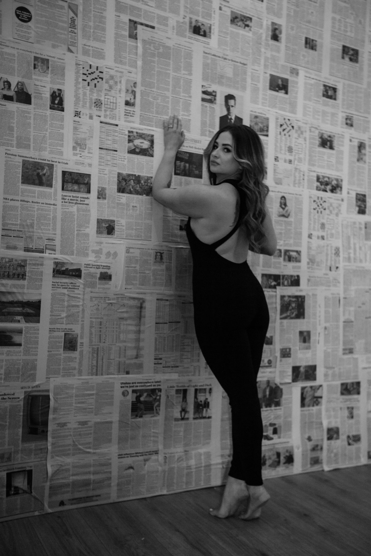 Person in black poses against a wall covered in newspaper clippings and photos.
