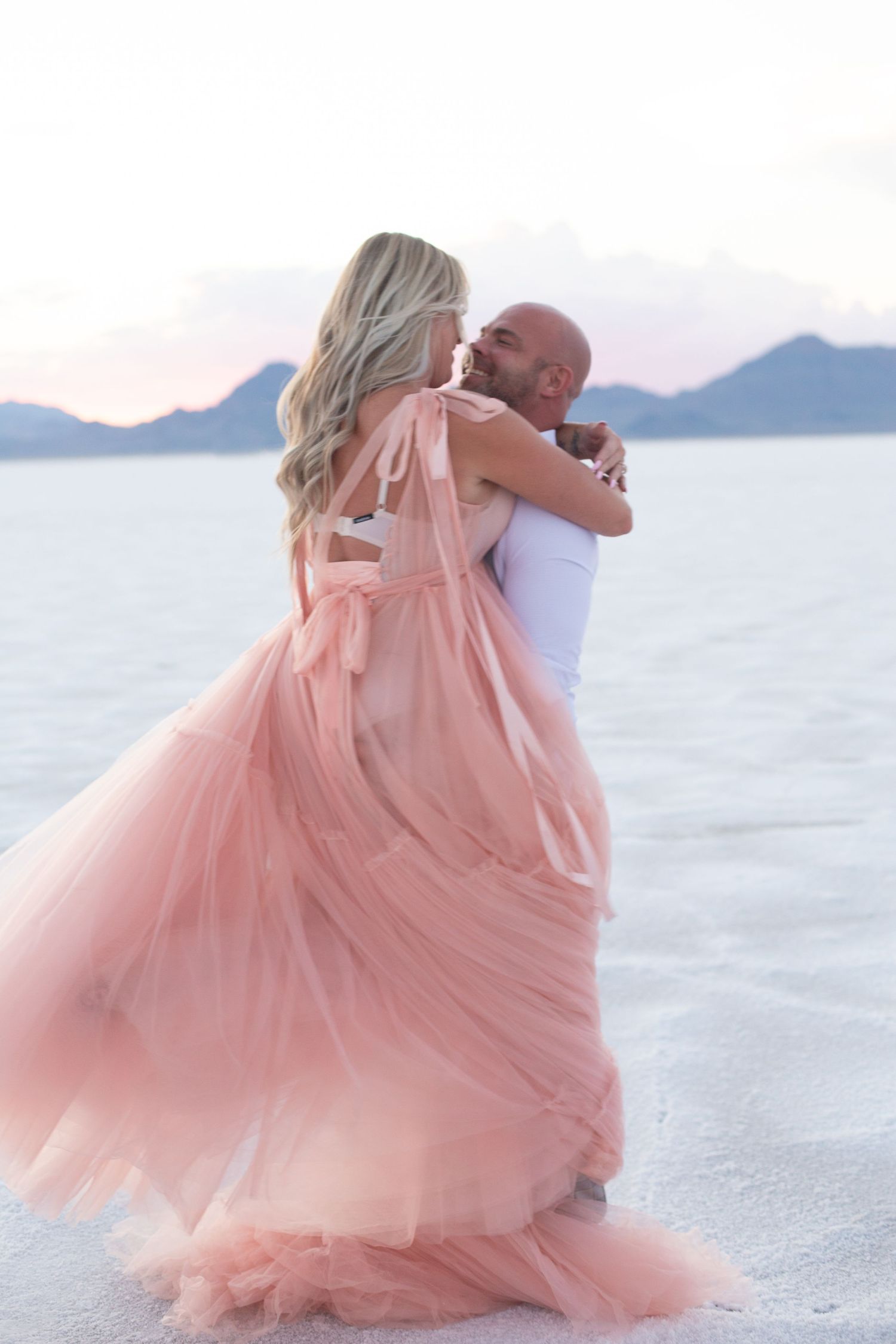 Romantic embrace on salt flats with flowing pink dress and mountain backdrop at sunset.