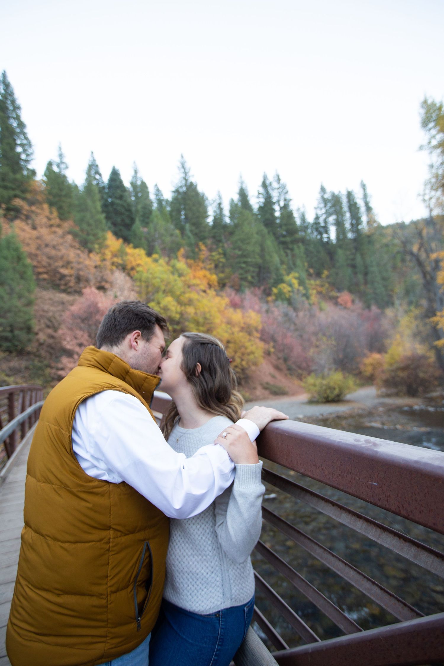 Couple sharing tender moment on wooden bench with autumn foliage in background.