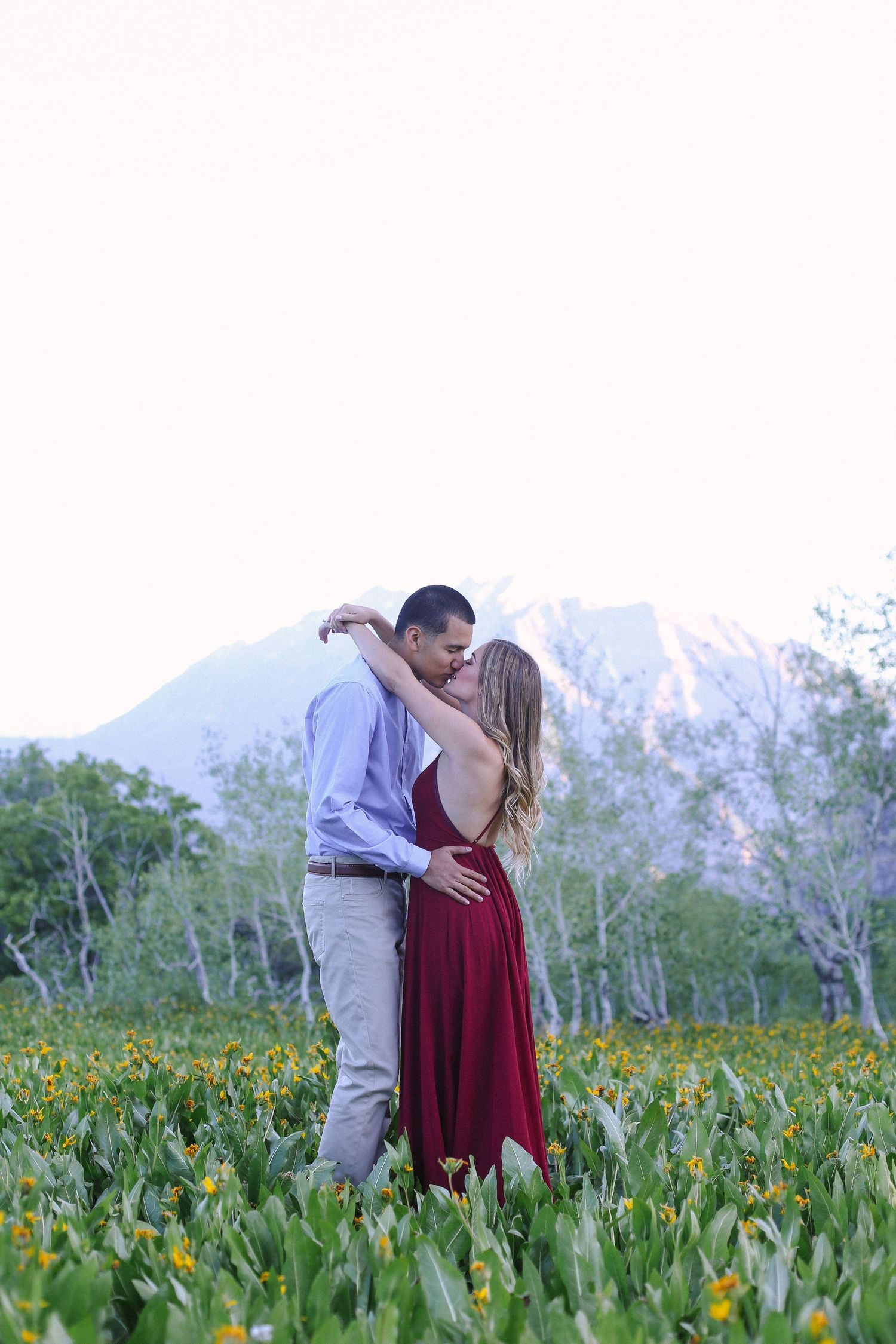 Couple sharing intimate moment in mountain wildflower meadow with snow-capped peaks in background.