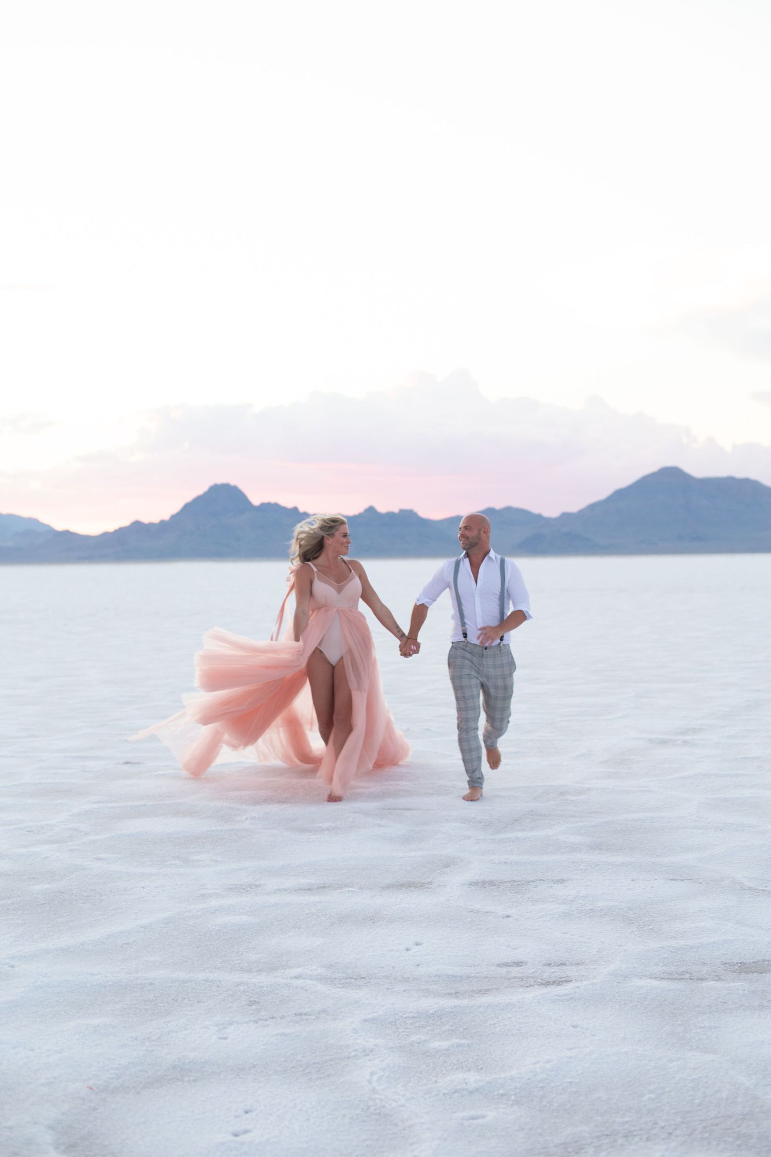 A couple runs through pristine white sand with mountains and pink skies in the background.