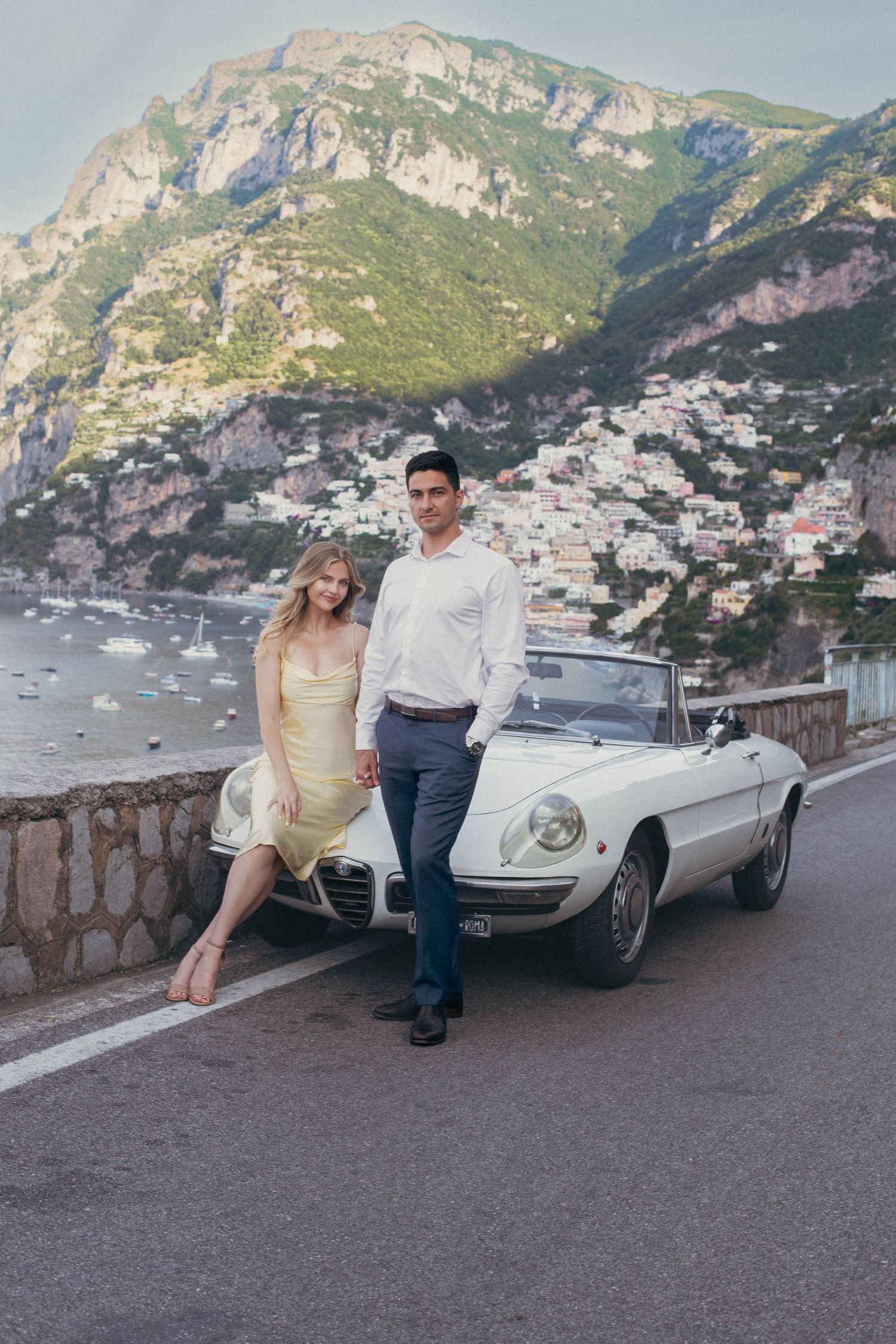 Elegant couple posing beside classic white Alfa Romeo Spider convertible on Amalfi Coast road in Positano.