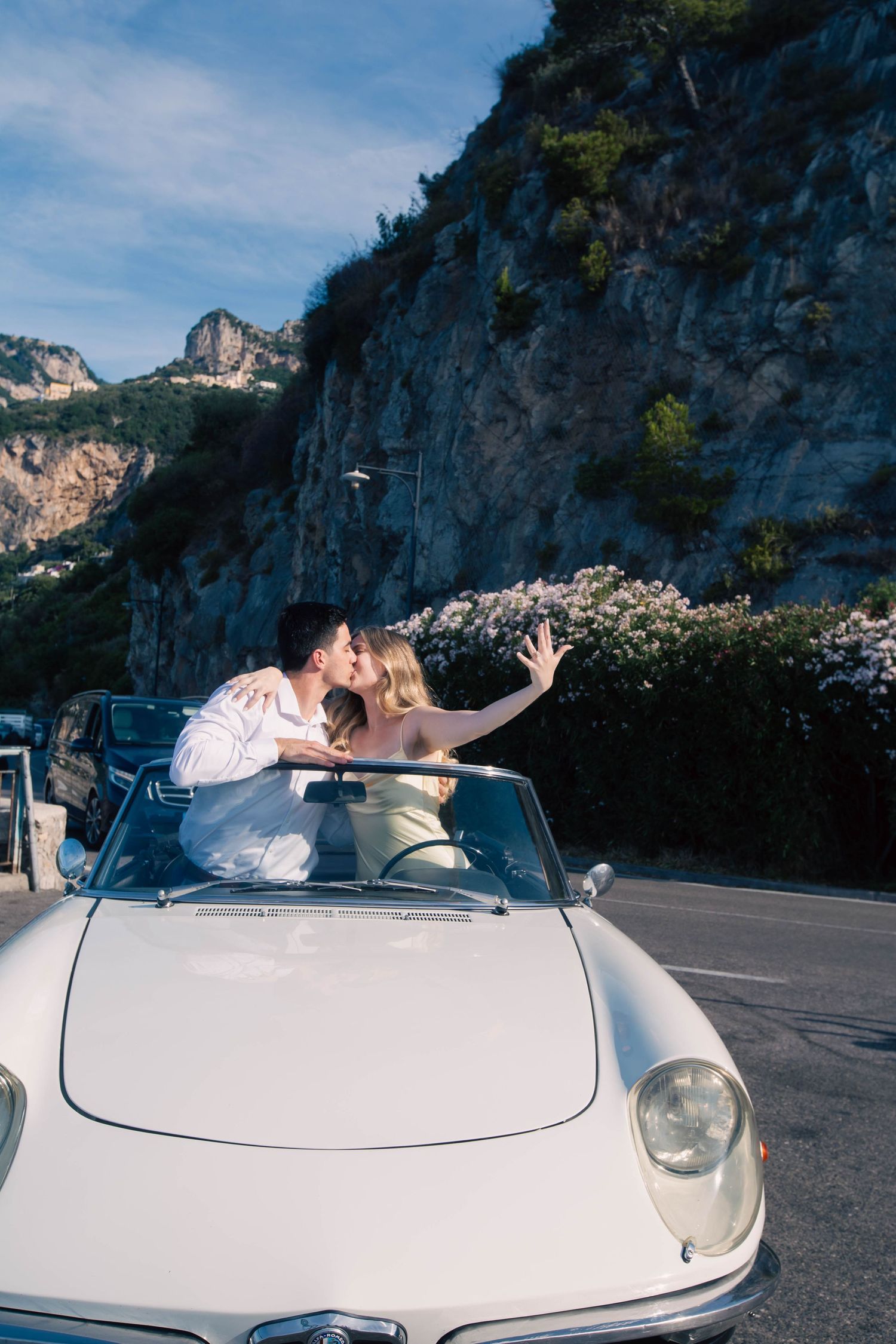 Couple kissing in classic white convertible car driving along scenic Amalfi Coast mountain road.