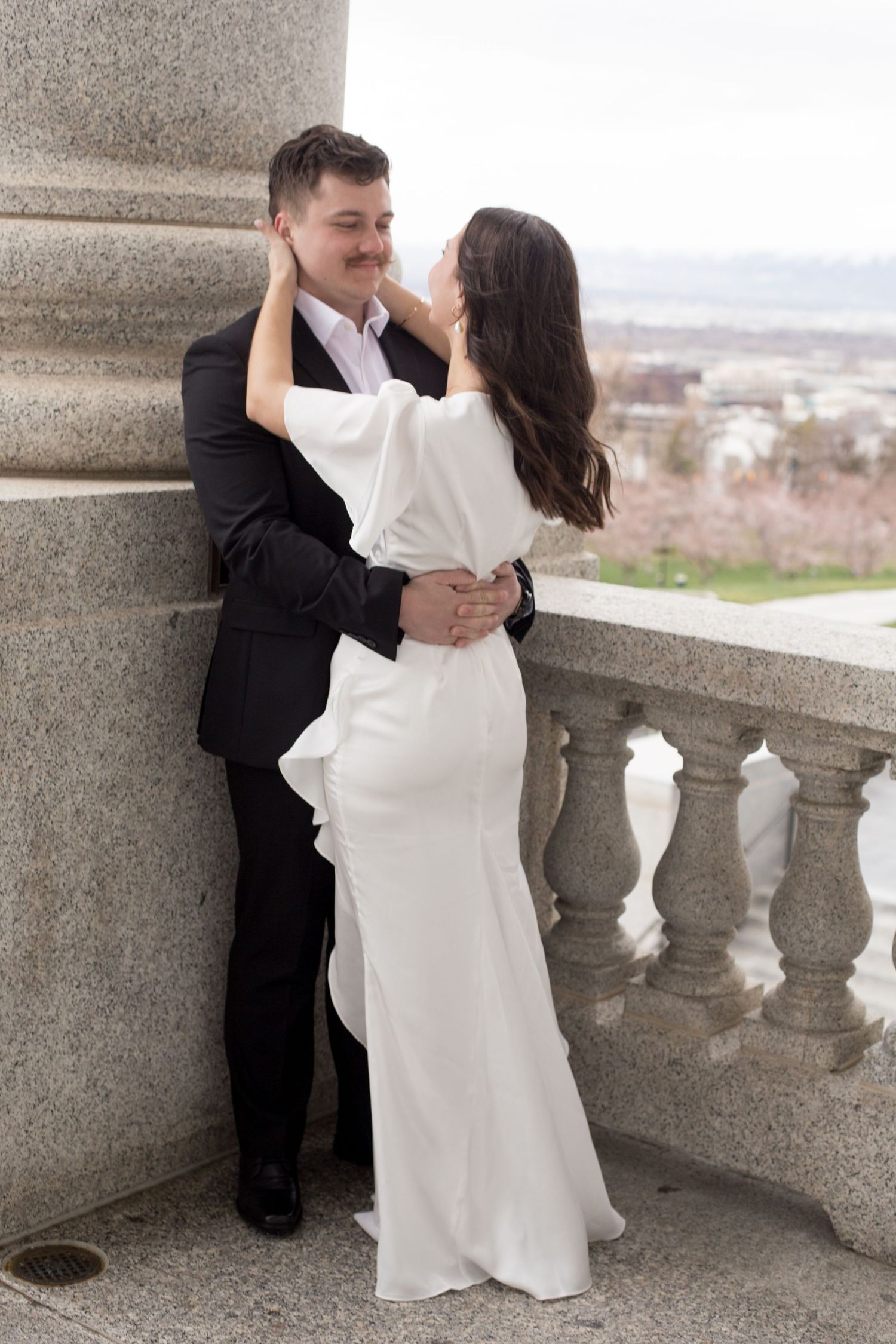 A romantic pose of newlyweds by stone columns with a cityscape view behind them.