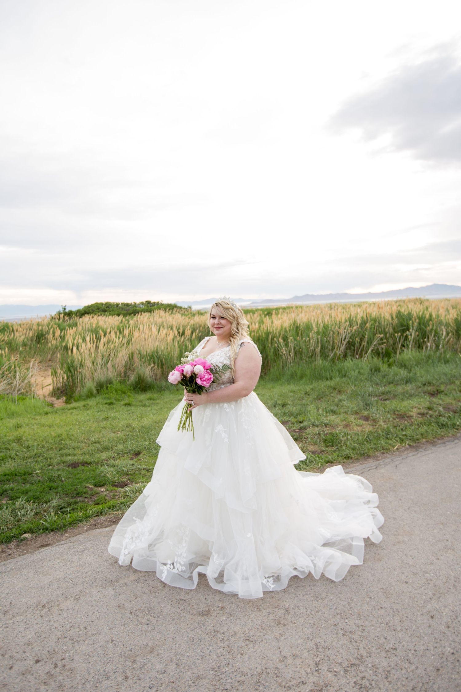A bride in a flowing white gown sits gracefully on a dirt path surrounded by rolling green hills.