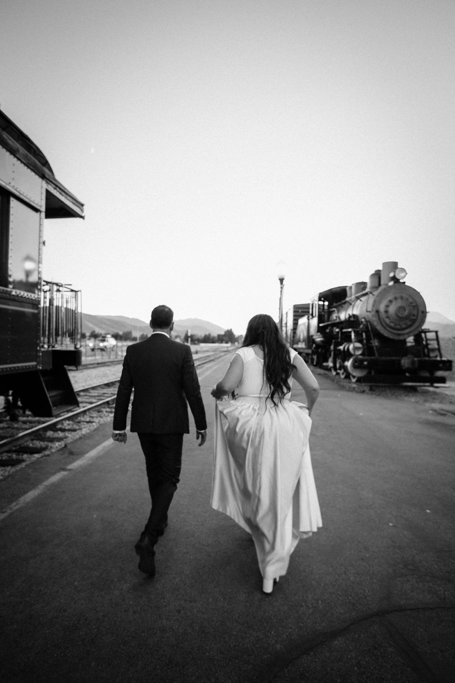 Black and white photo of couple walking along train tracks in wedding attire.