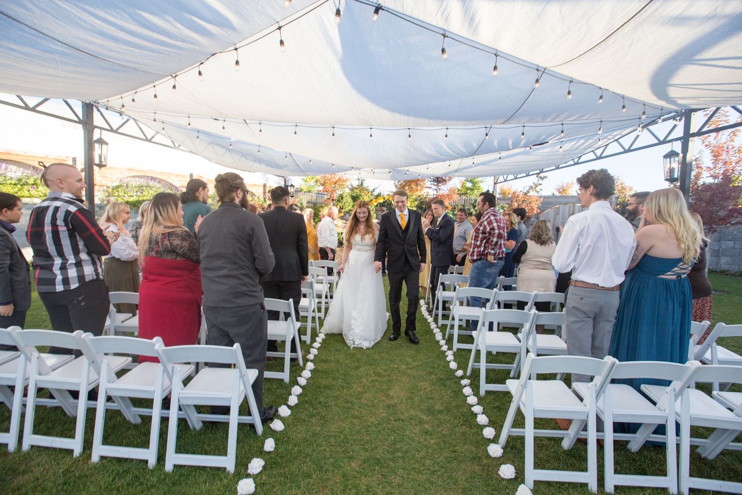 Outdoor wedding ceremony under a white tent with guests standing as bride and groom walk down the aisle lined with white chairs.