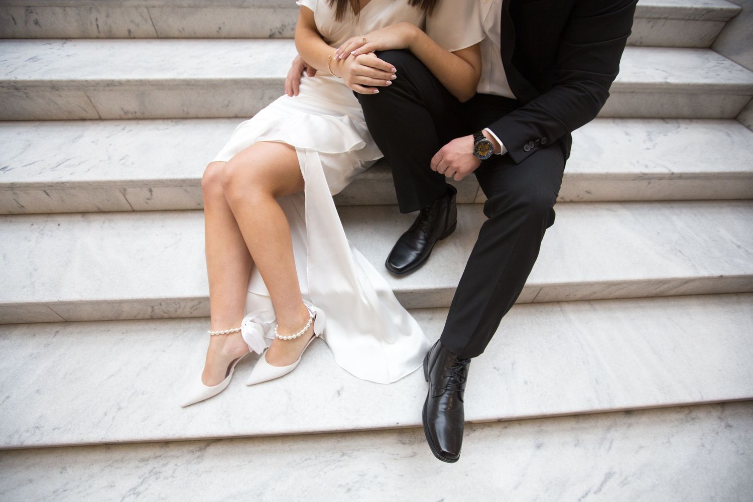 Couple sitting on marble steps, woman in white dress with silver heels, man in black suit with dress shoes.