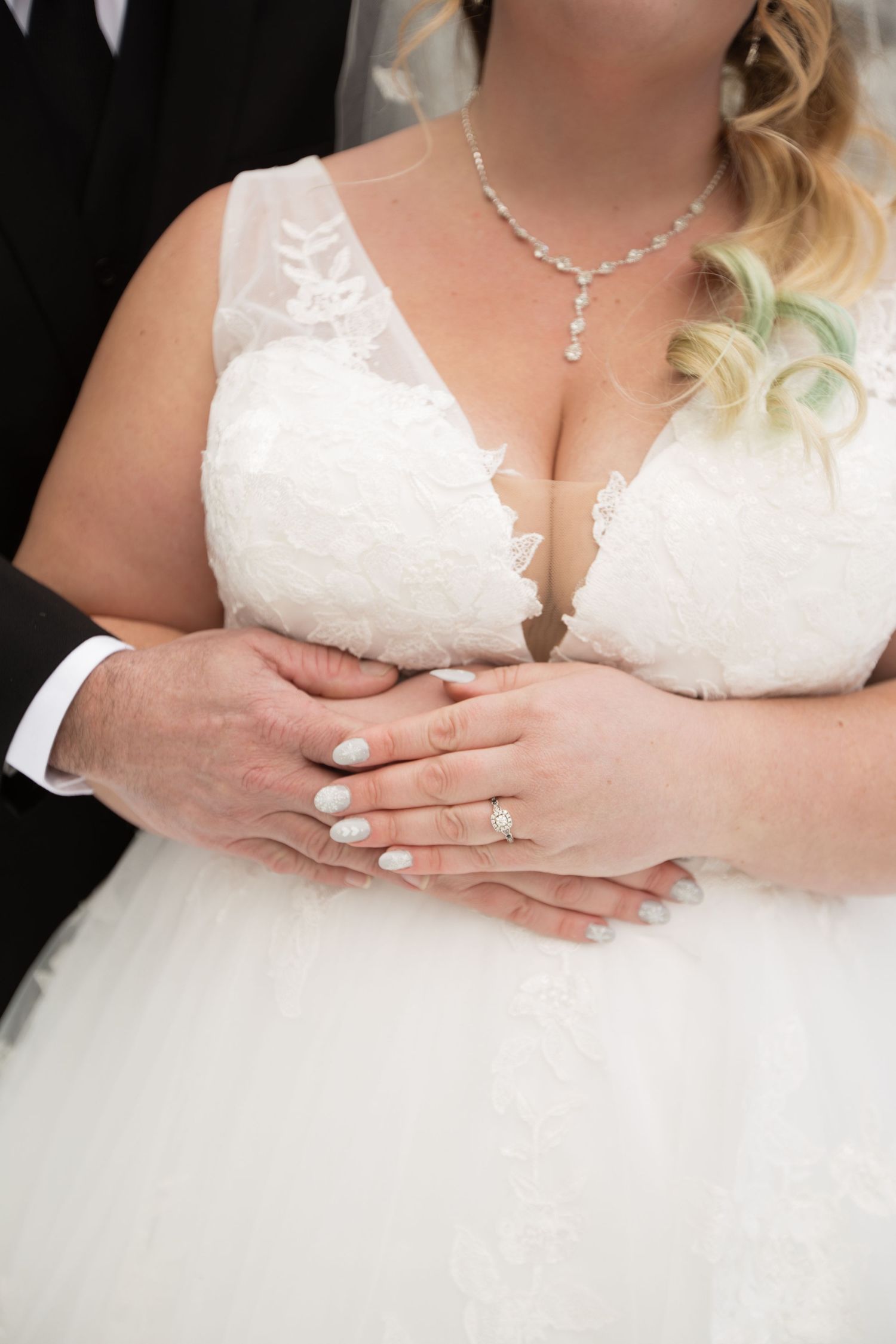 Close-up details of a white lace wedding dress with delicate necklace and hands clasped.