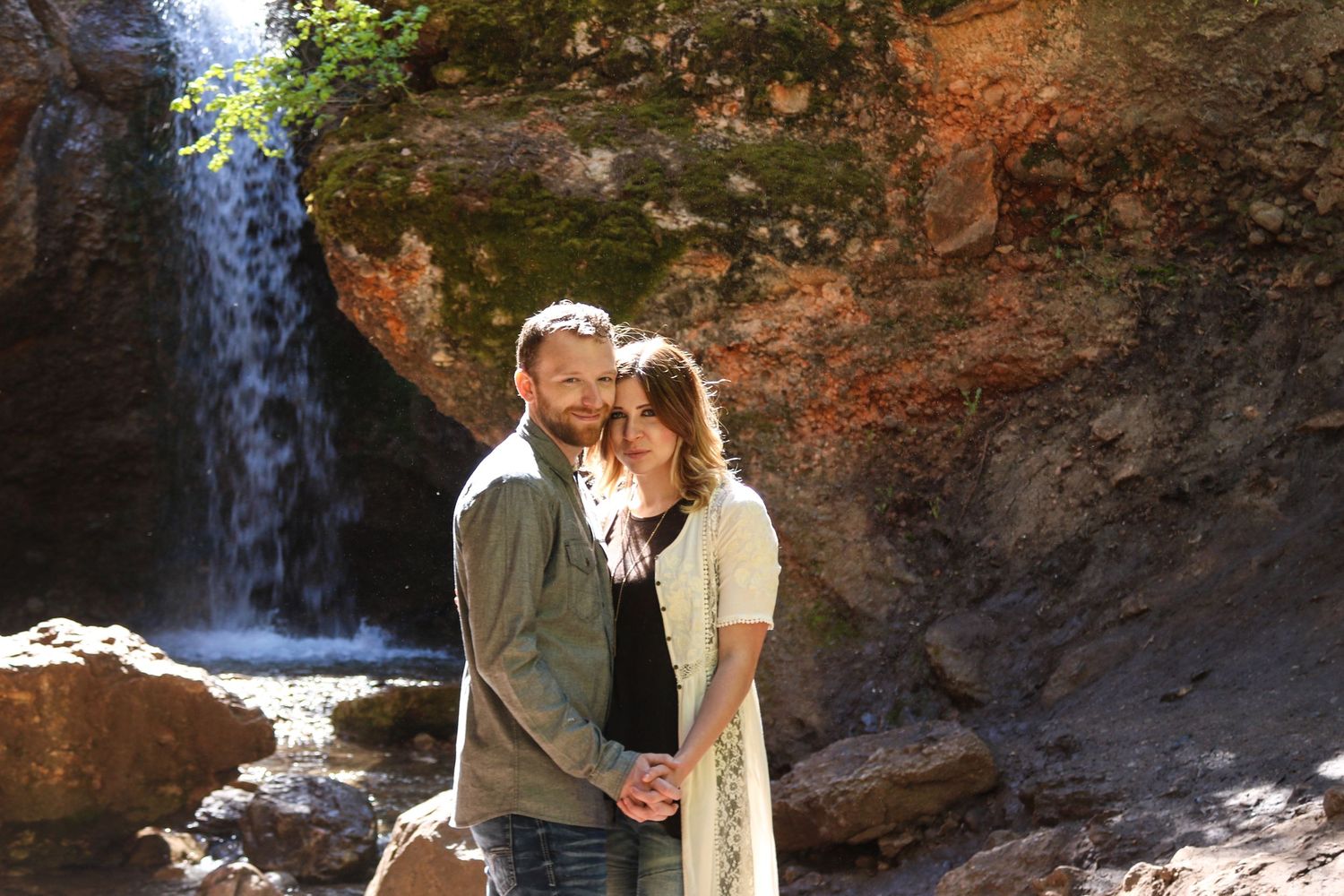 A couple shares an intimate moment in front of a scenic waterfall among rocky cliffs.