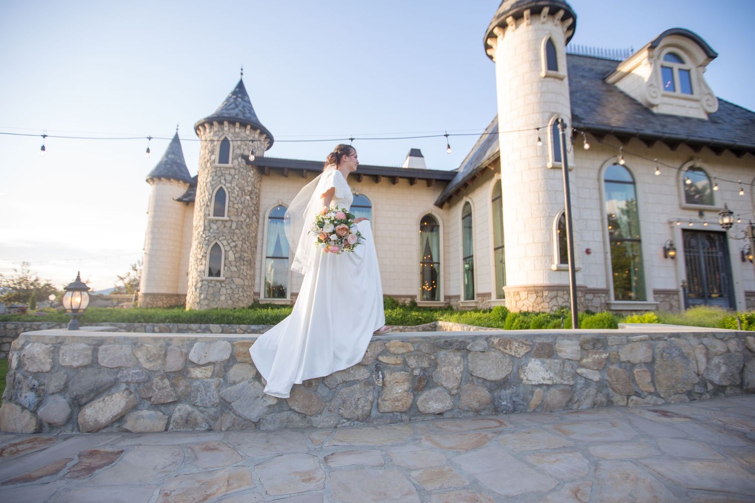 Bride in white dress standing outside a castle-like venue with stone walls and turrets.