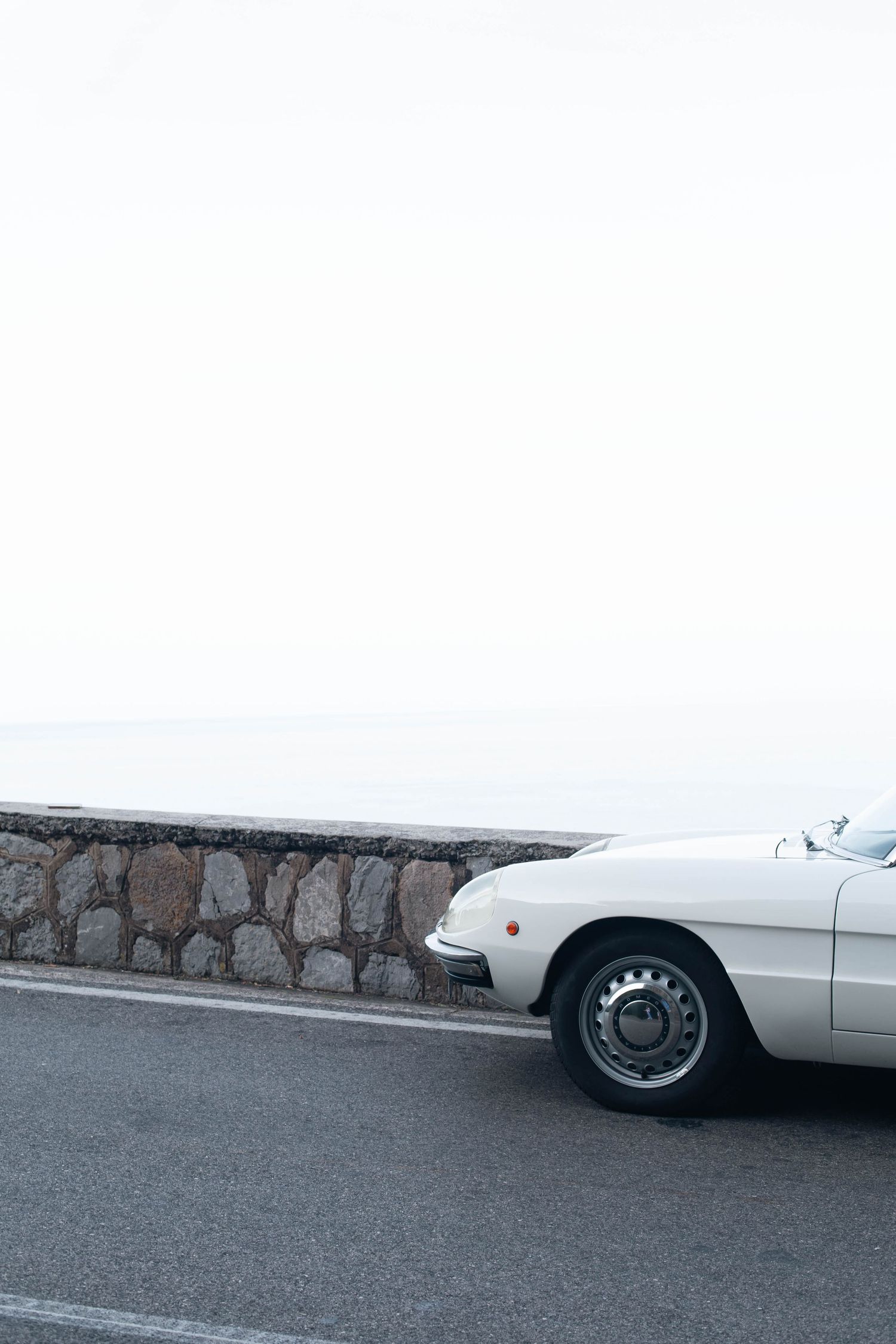 A classic white vintage sports car parked on a coastal road beside a stone wall with a misty ocean backdrop.