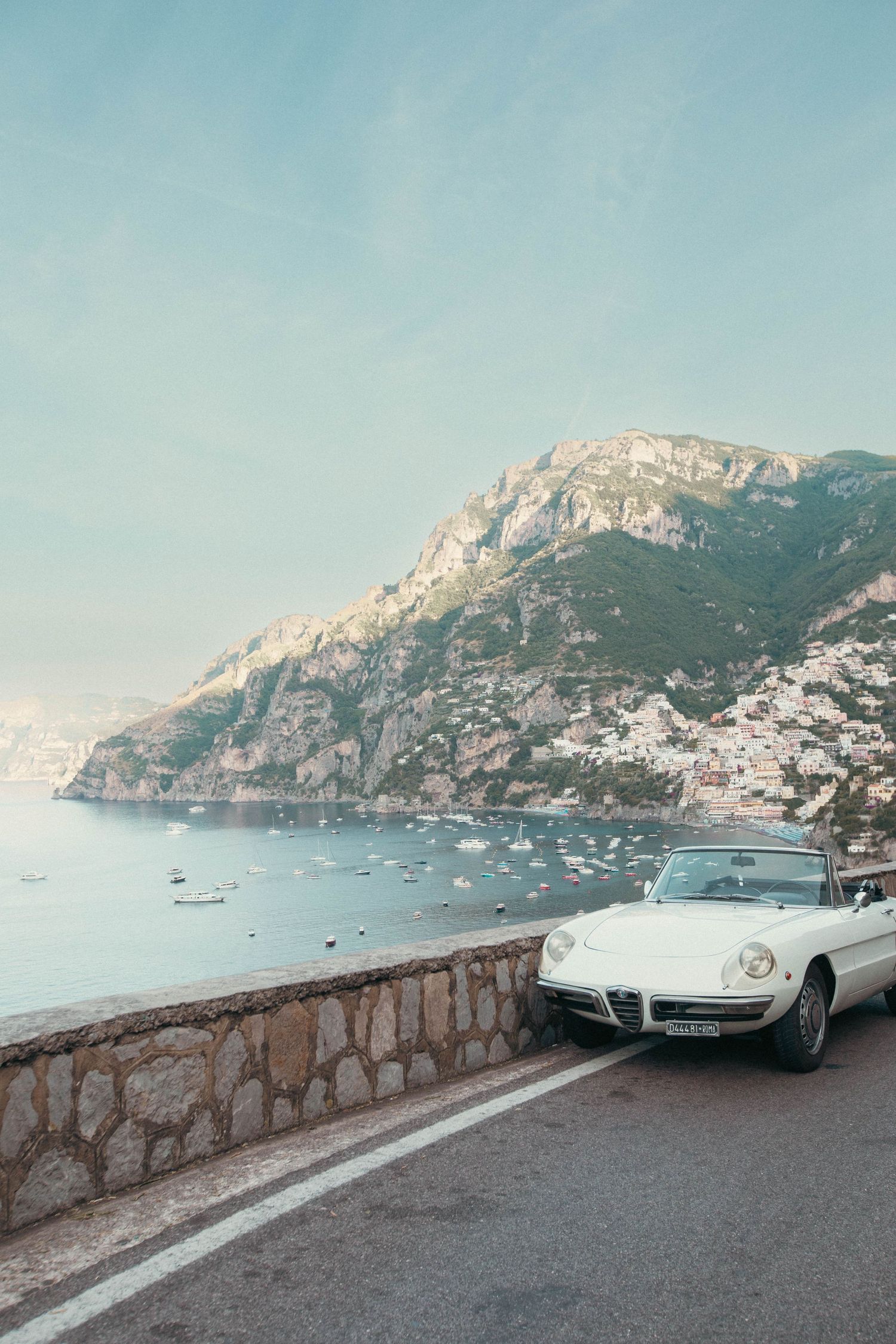 White vintage convertible parked on Amalfi Coast road with stunning sea and cliffside village views.