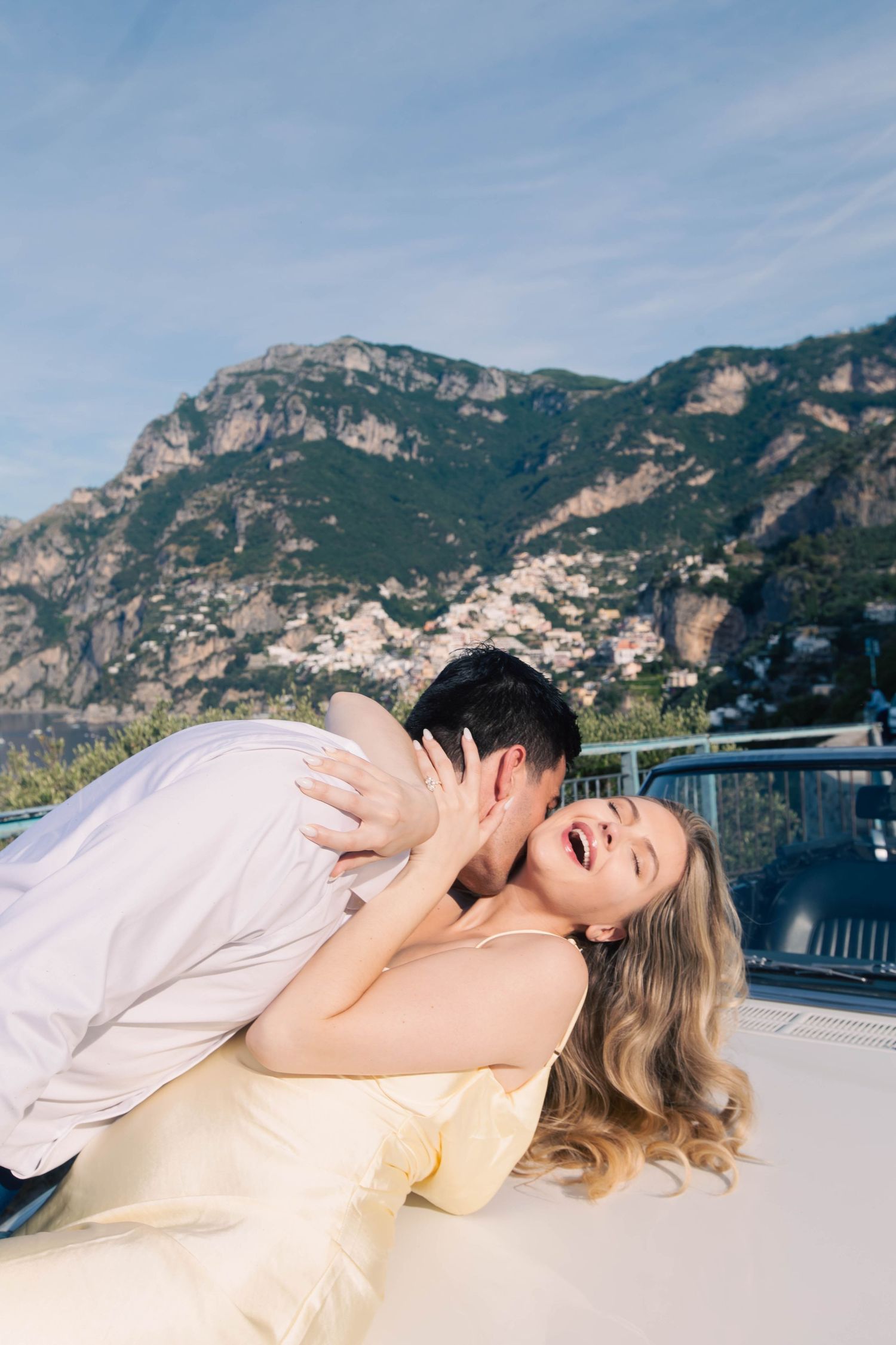 Joyful couple sharing a playful moment on a classic car hood with scenic Amalfi Coast mountains behind them.