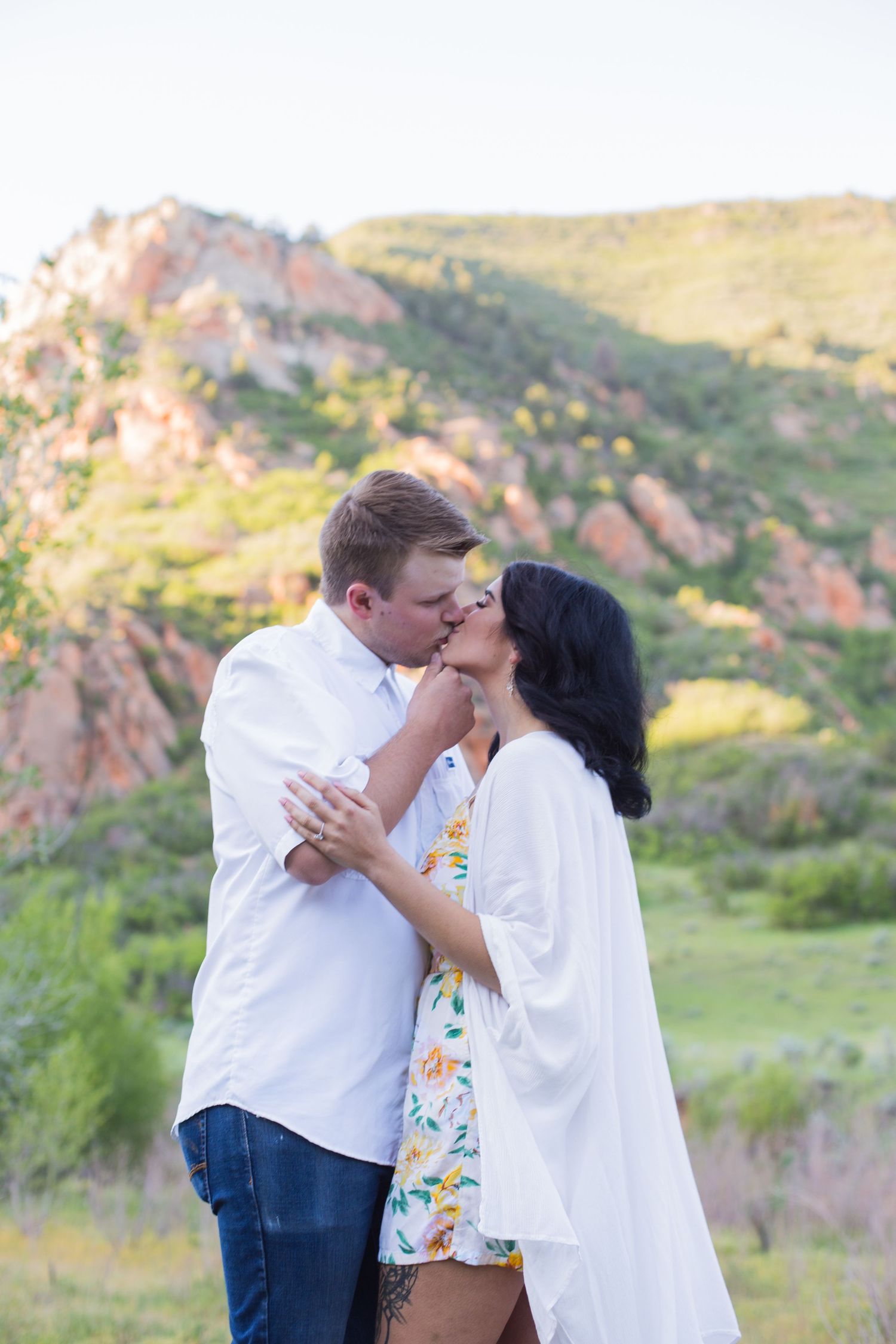 Couple sharing a romantic kiss outdoors with scenic rocky mountain landscape and green hills in background.
