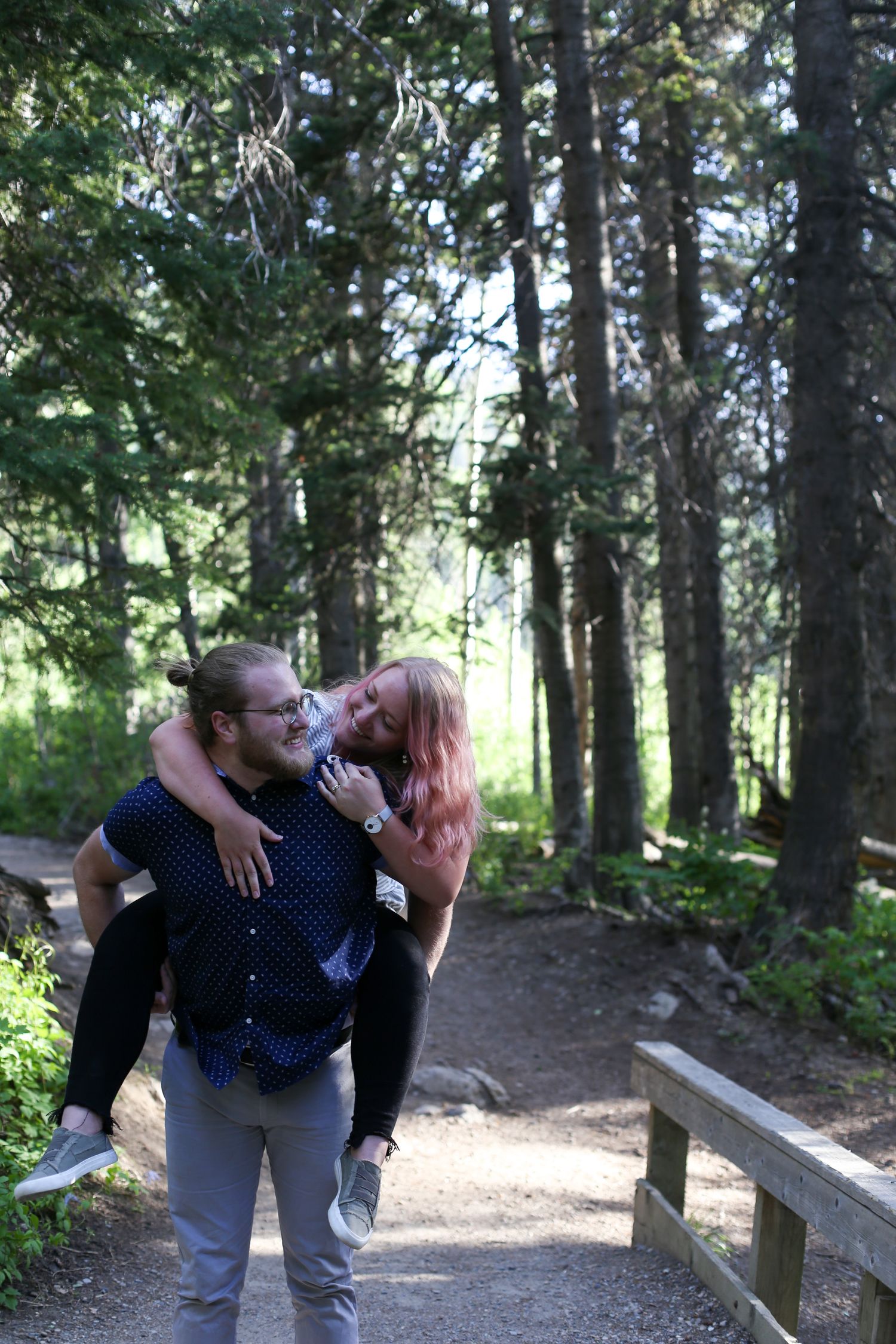 A man gives a woman a piggyback ride on a forest trail, both smiling and enjoying a sunny outdoor adventure.