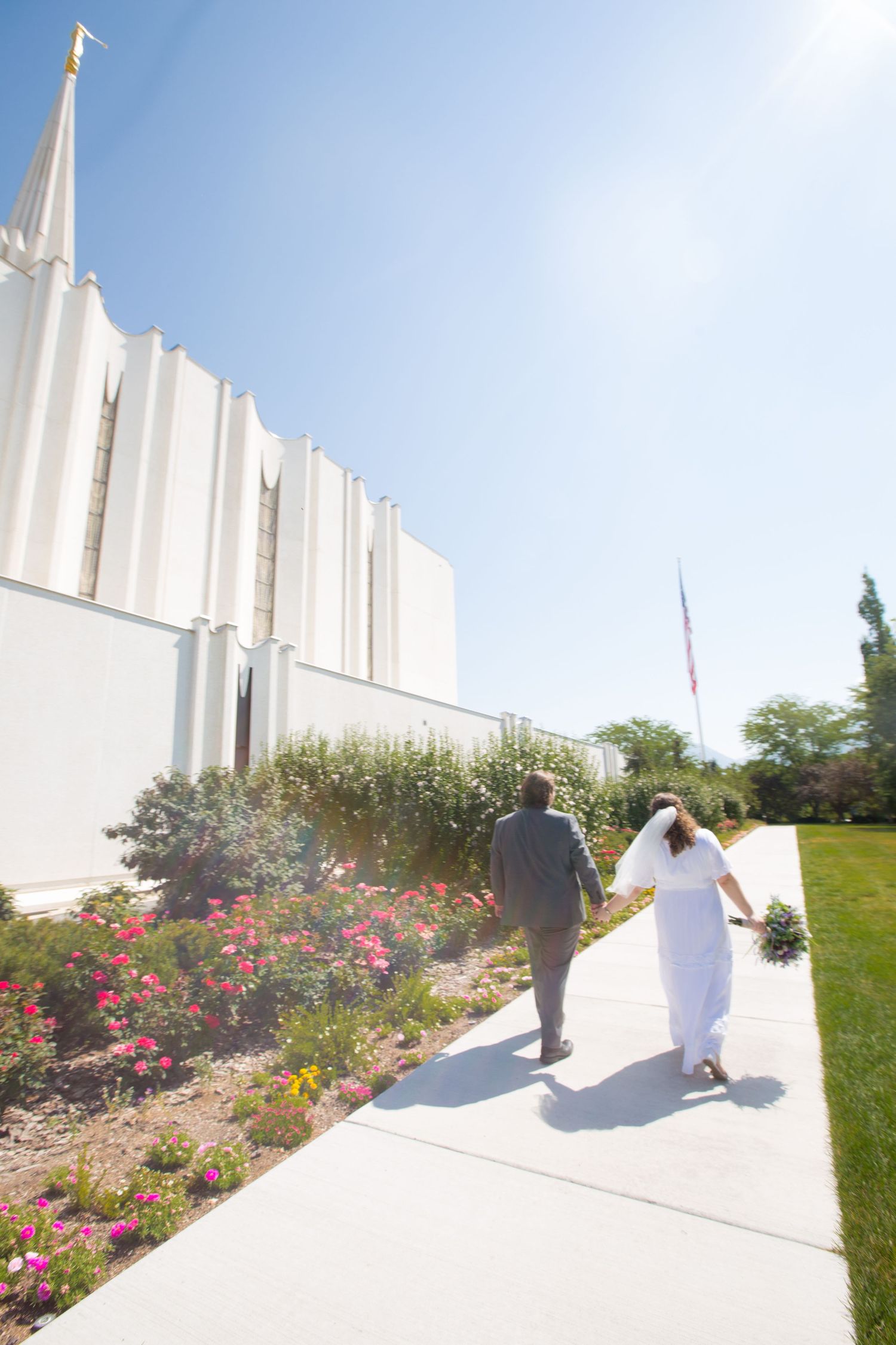 Bride and groom walking hand in hand along a sunlit path beside a white LDS temple with colorful flowers.