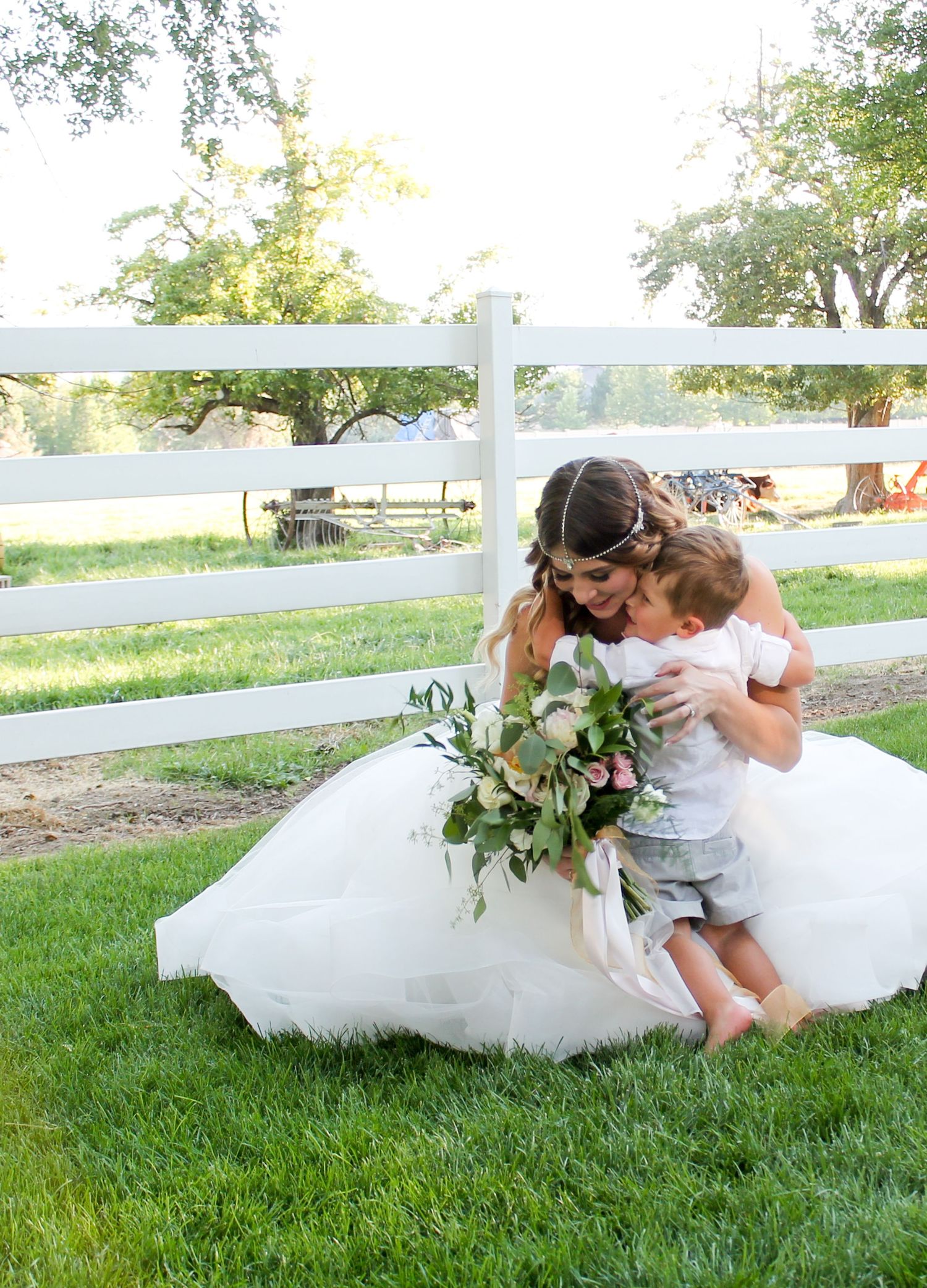 Bride in white tulle gown kneeling on green grass hugging a toddler while holding a lush floral bouquet near white fence.