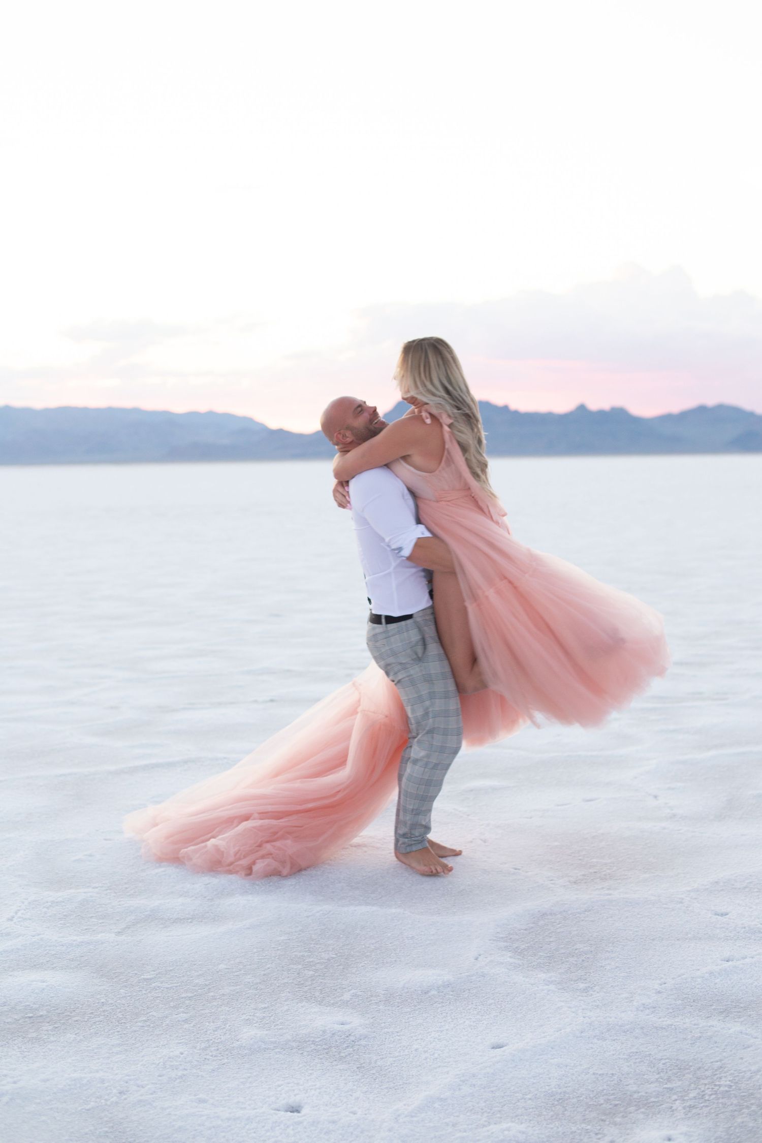Dancing couple on salt flats with pink flowing dress against purple mountain sunset.