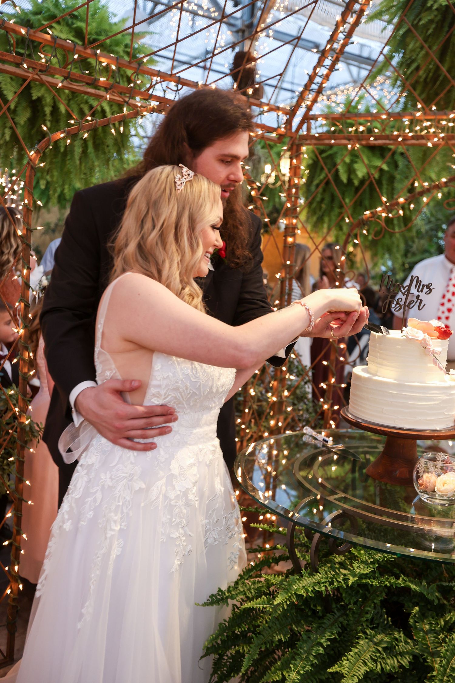 Wedding cake cutting moment under string lights and greenery in greenhouse setting.