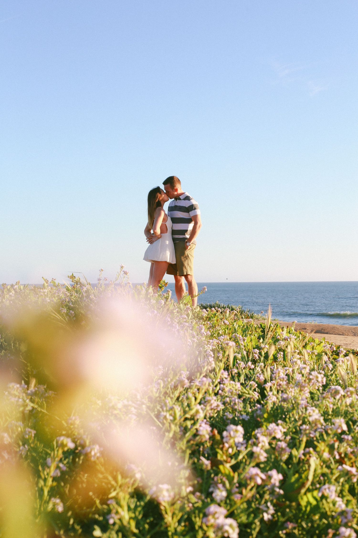 Couple embraces in a field of wildflowers with ocean views at sunset.