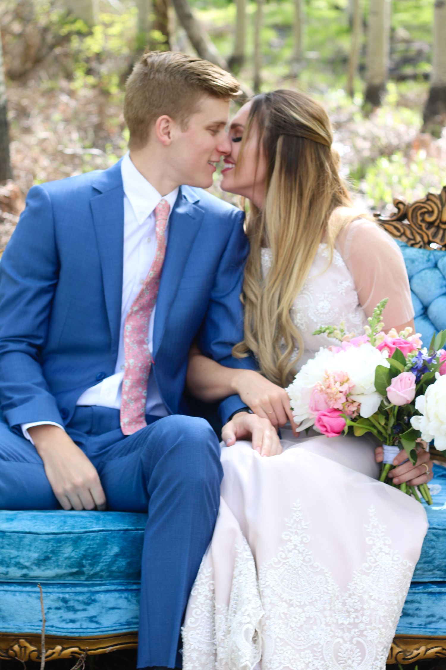 Romantic wedding couple sitting on blue vintage couch in outdoor garden setting with pink and white floral bouquet.