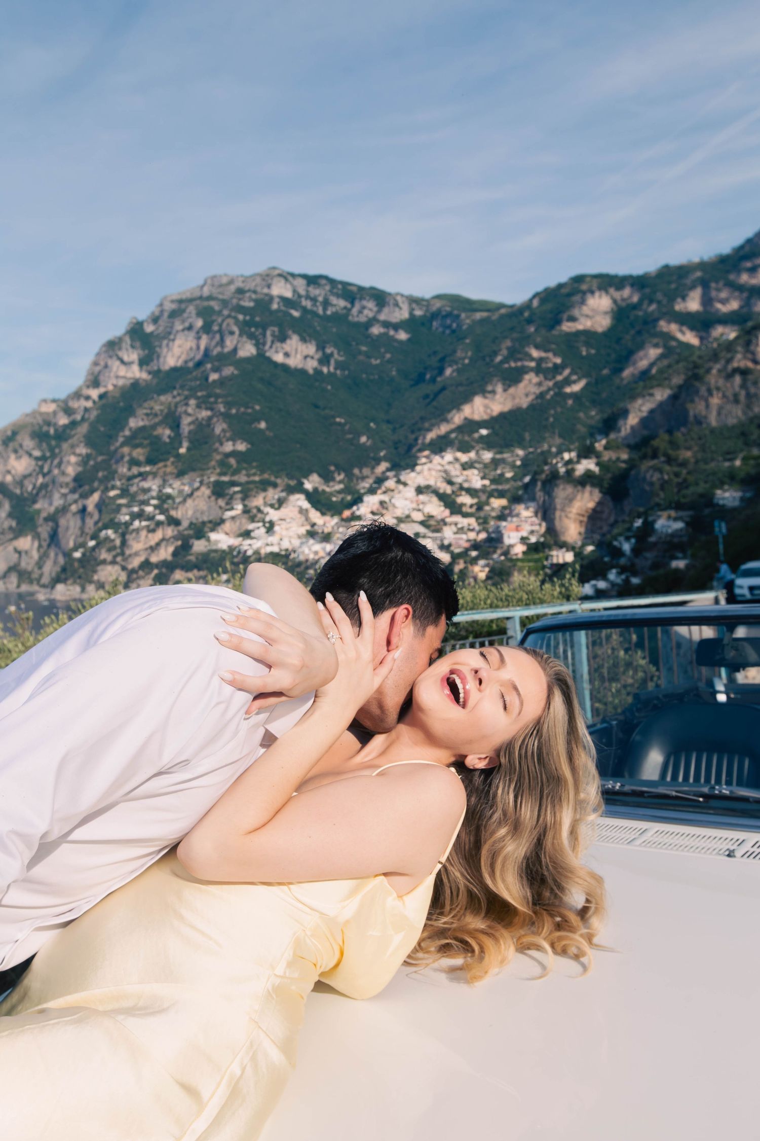 Joyful couple in playful romantic moment on white vintage car with Positano mountain village behind them.