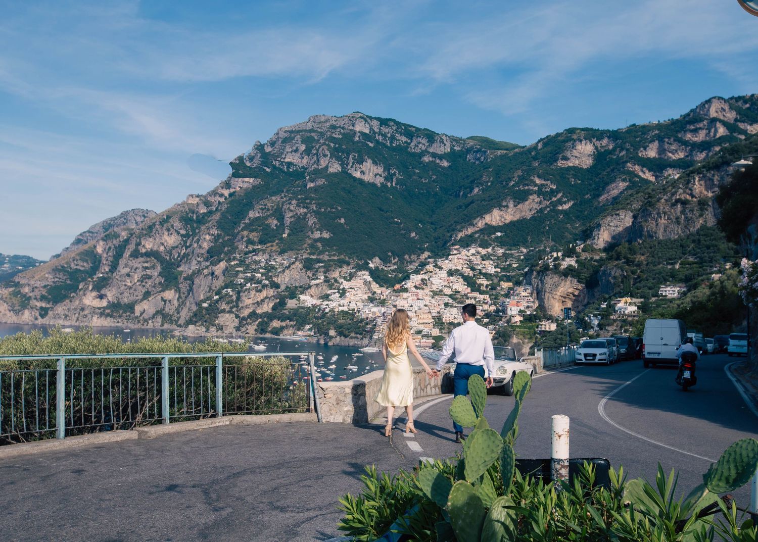 Couple walking along scenic Amalfi Coast road overlooking Positano village and lush mountain landscape in Italy.